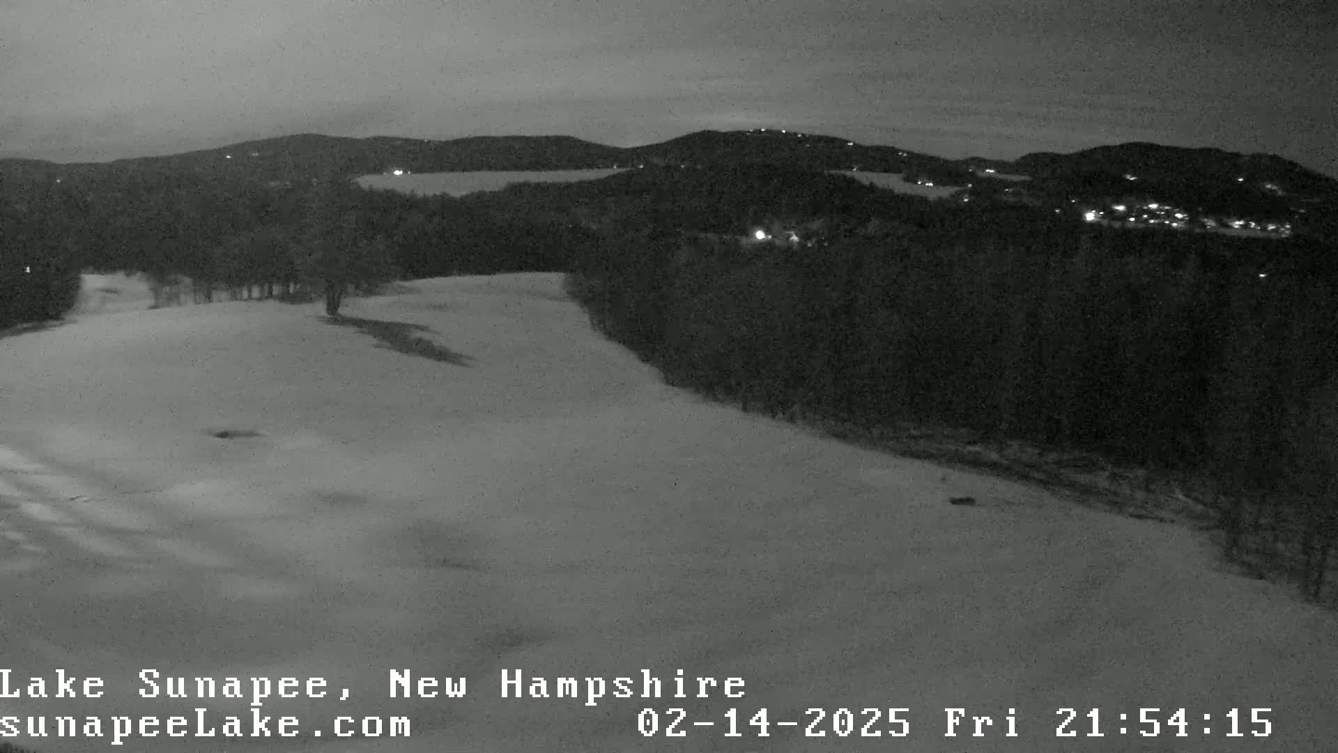 A snow-covered landscape at night, showing a hill sloping down toward a dark forest and distant lights from a town beyond.