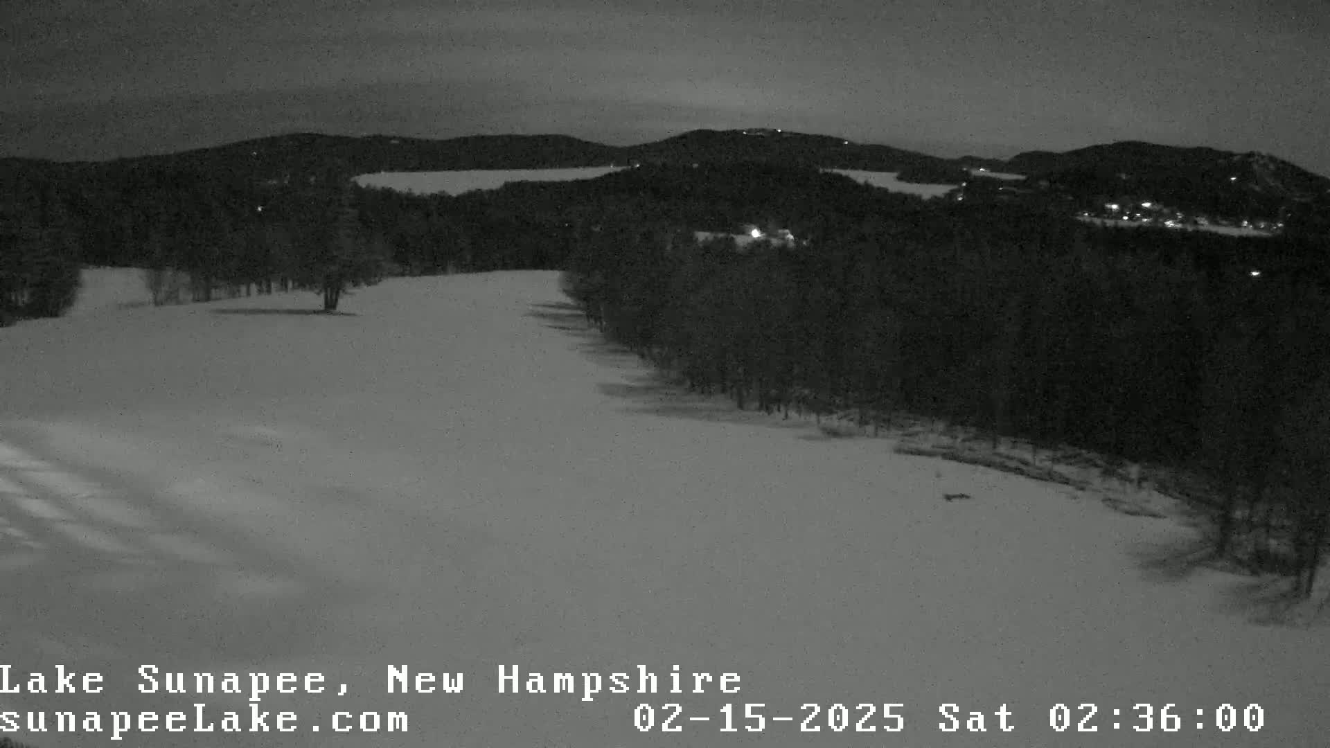 A snow-covered landscape at night, featuring a dark forest and a body of water in the distance under a dusky sky.