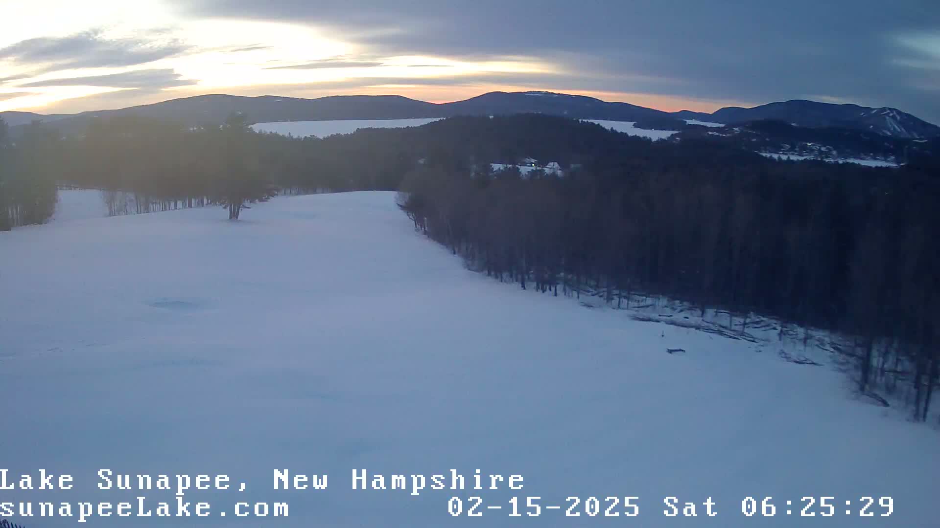 A snow-covered landscape at sunset, featuring a line of trees bordering a wide, open snowy area, with mountains and a lake visible in the distance.