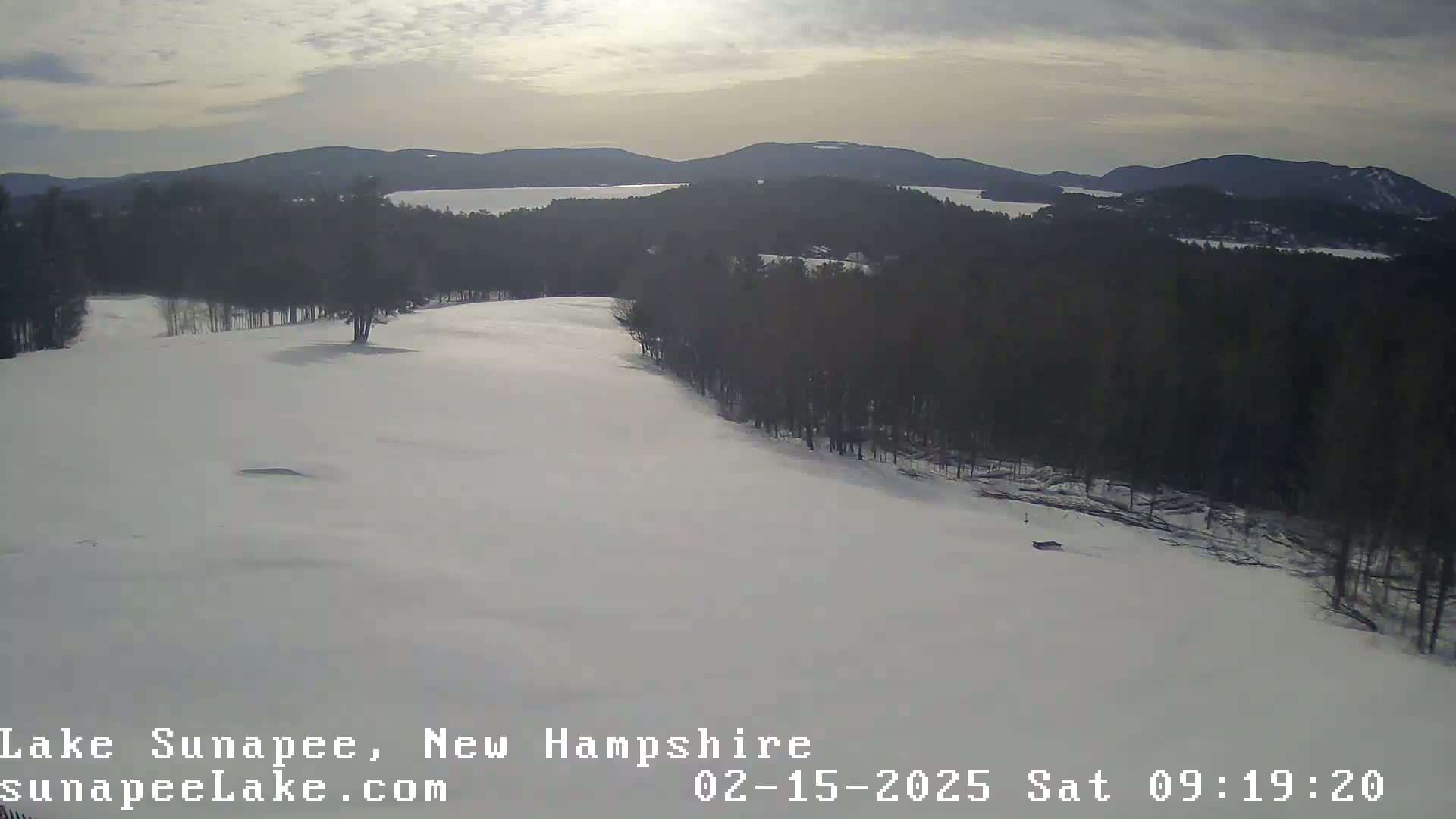A snow-covered landscape with a line of trees separating a large expanse of snow from a distant lake and mountains under a partly cloudy sky.