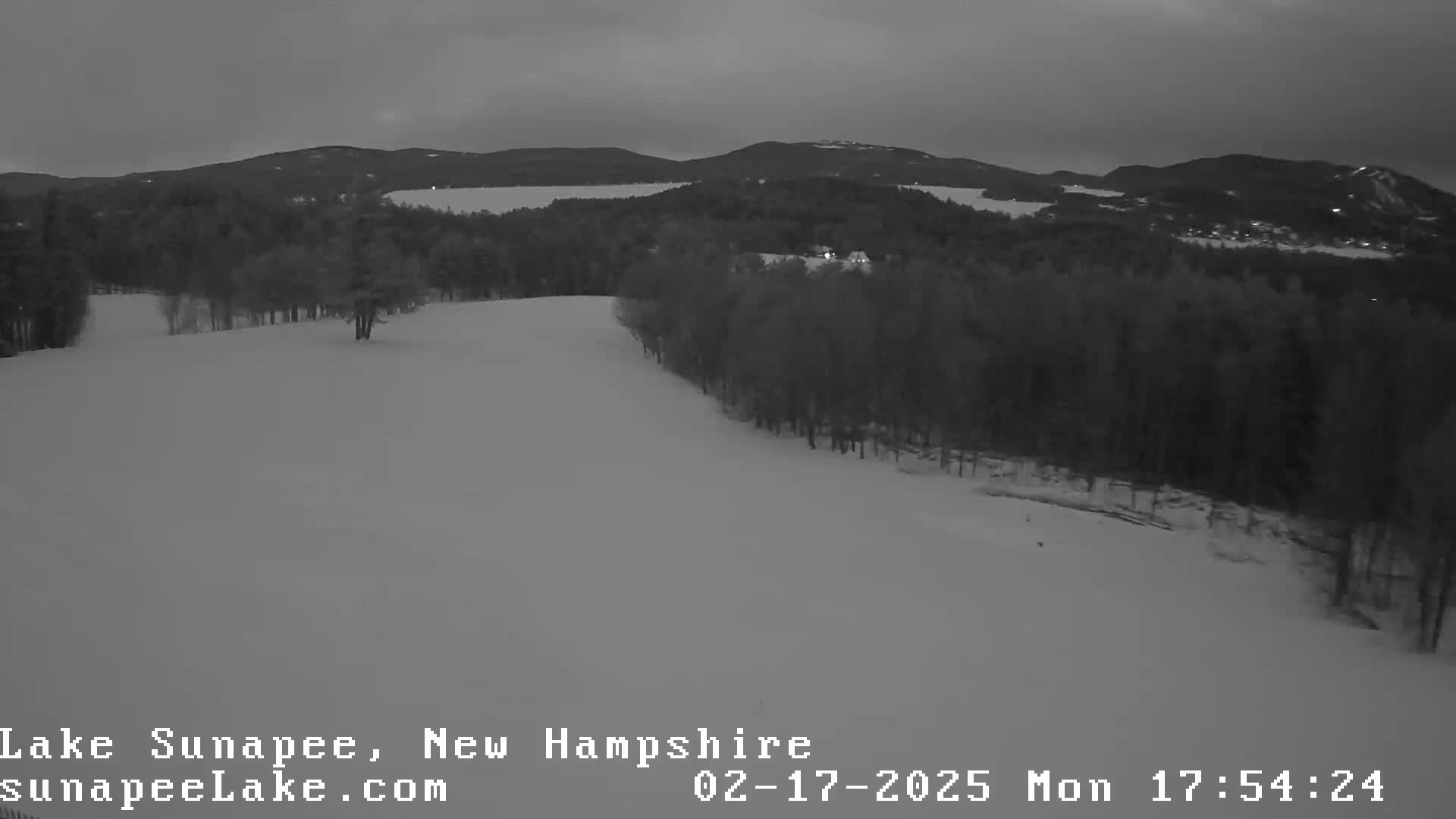 A snow-covered landscape with dark, wooded hills in the background under a cloudy, twilight sky.