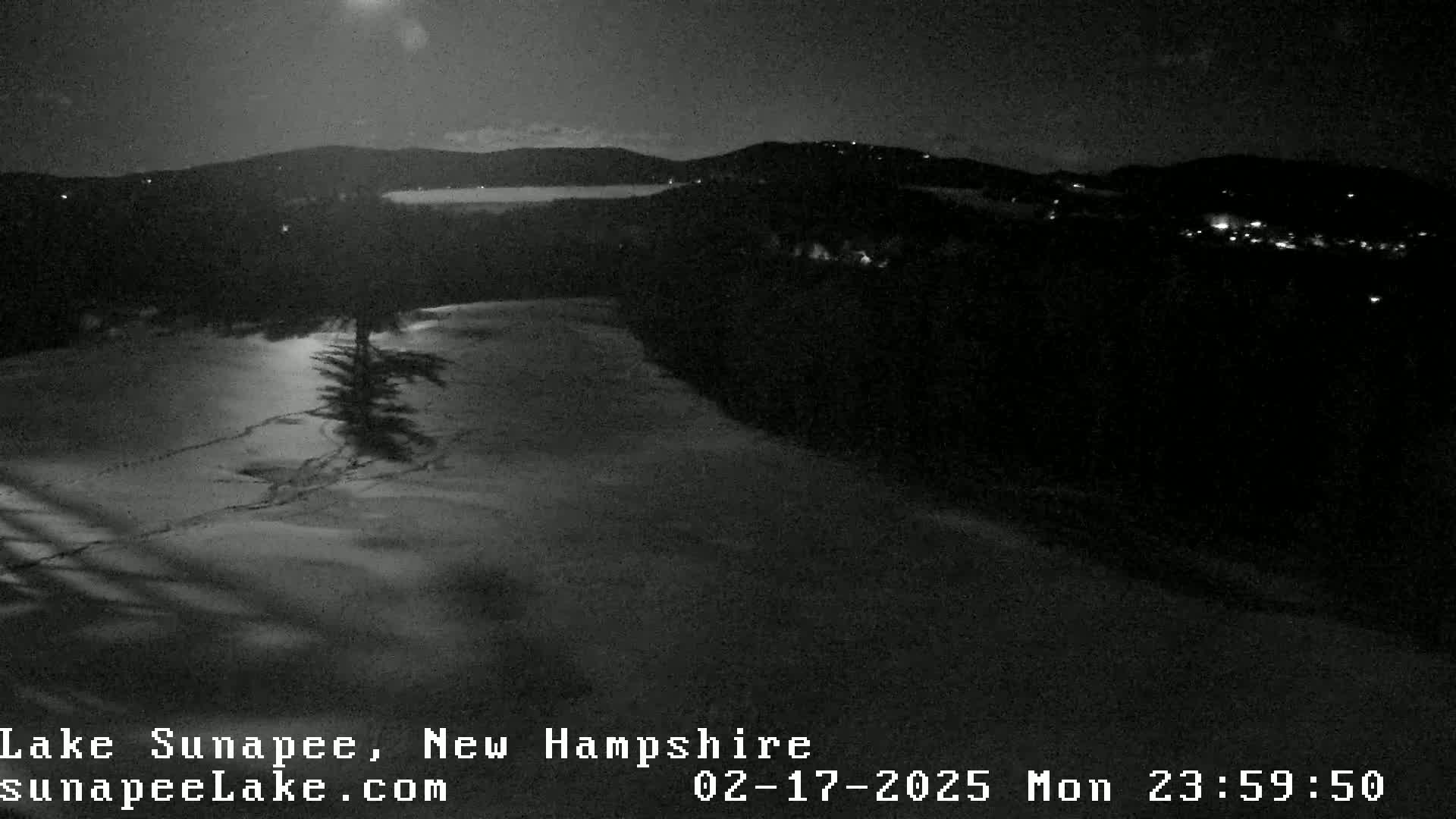 A nighttime, snow-covered landscape shows a dark, wooded area and a body of water in the distance under a mostly clear sky.