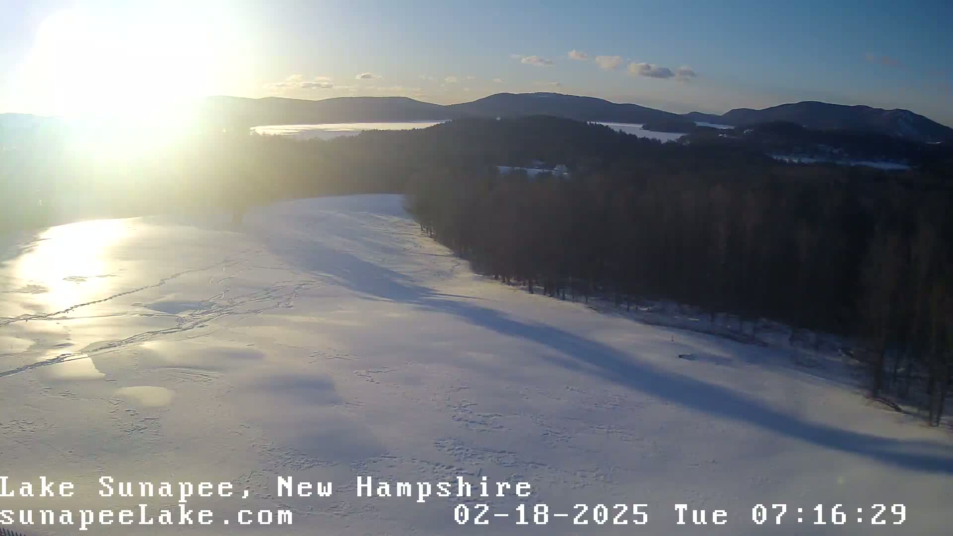 A bright, sunny day shows a frozen lake, partially covered in snow, with a dark evergreen forest in the background, and distant mountains under a mostly clear sky.
