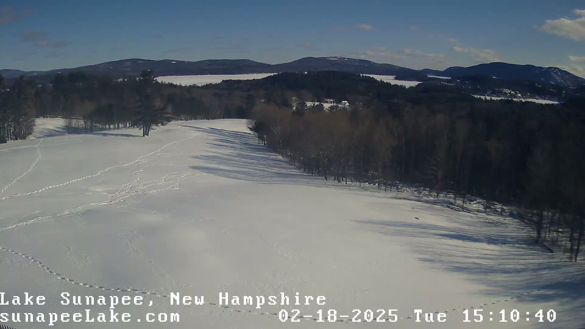 A snow-covered field with footprints and a line of trees leading to a frozen lake and distant mountains under a clear blue sky.