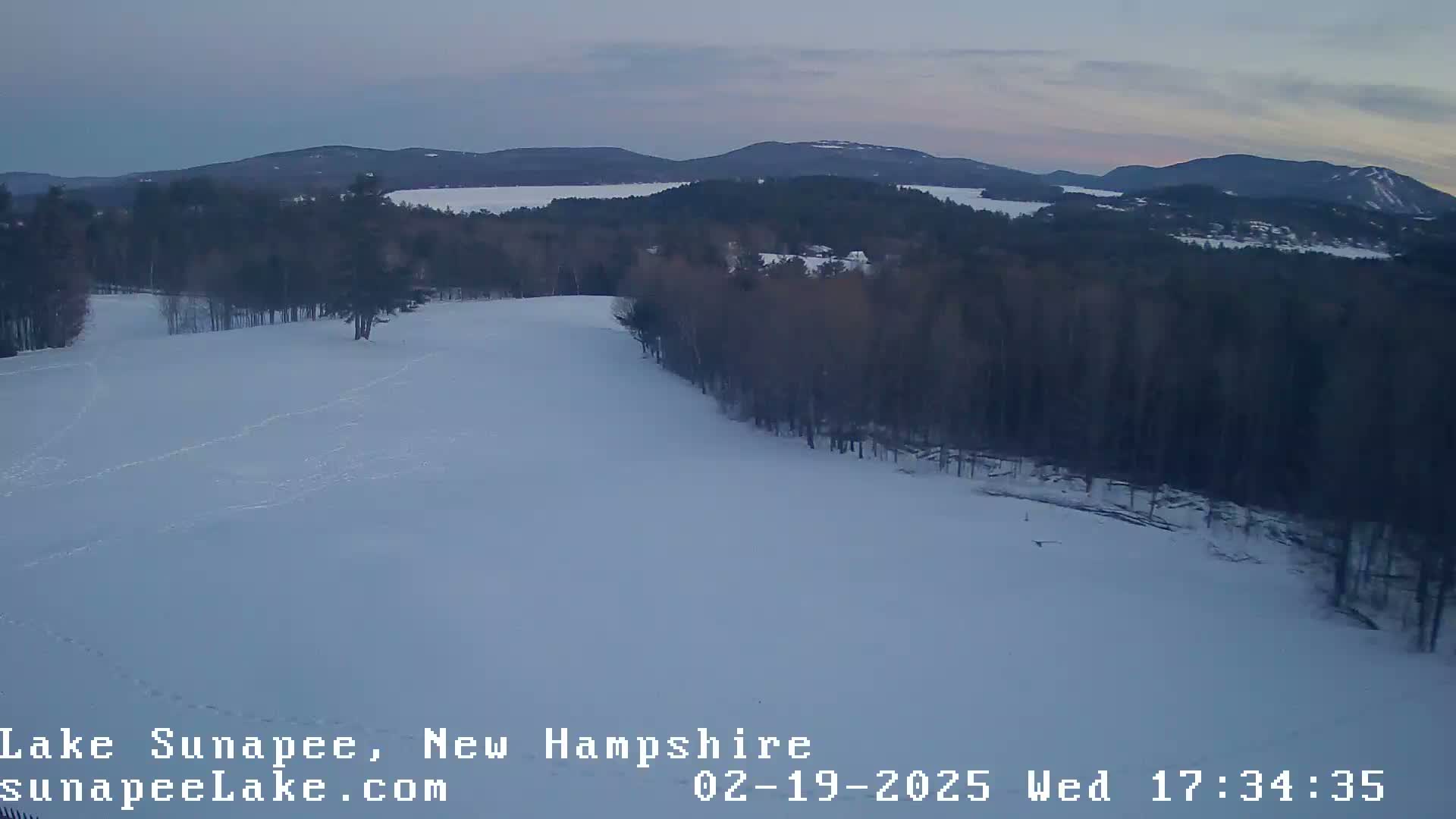 A snow-covered landscape at dusk shows a hilly area with dark trees and a lake in the distance.