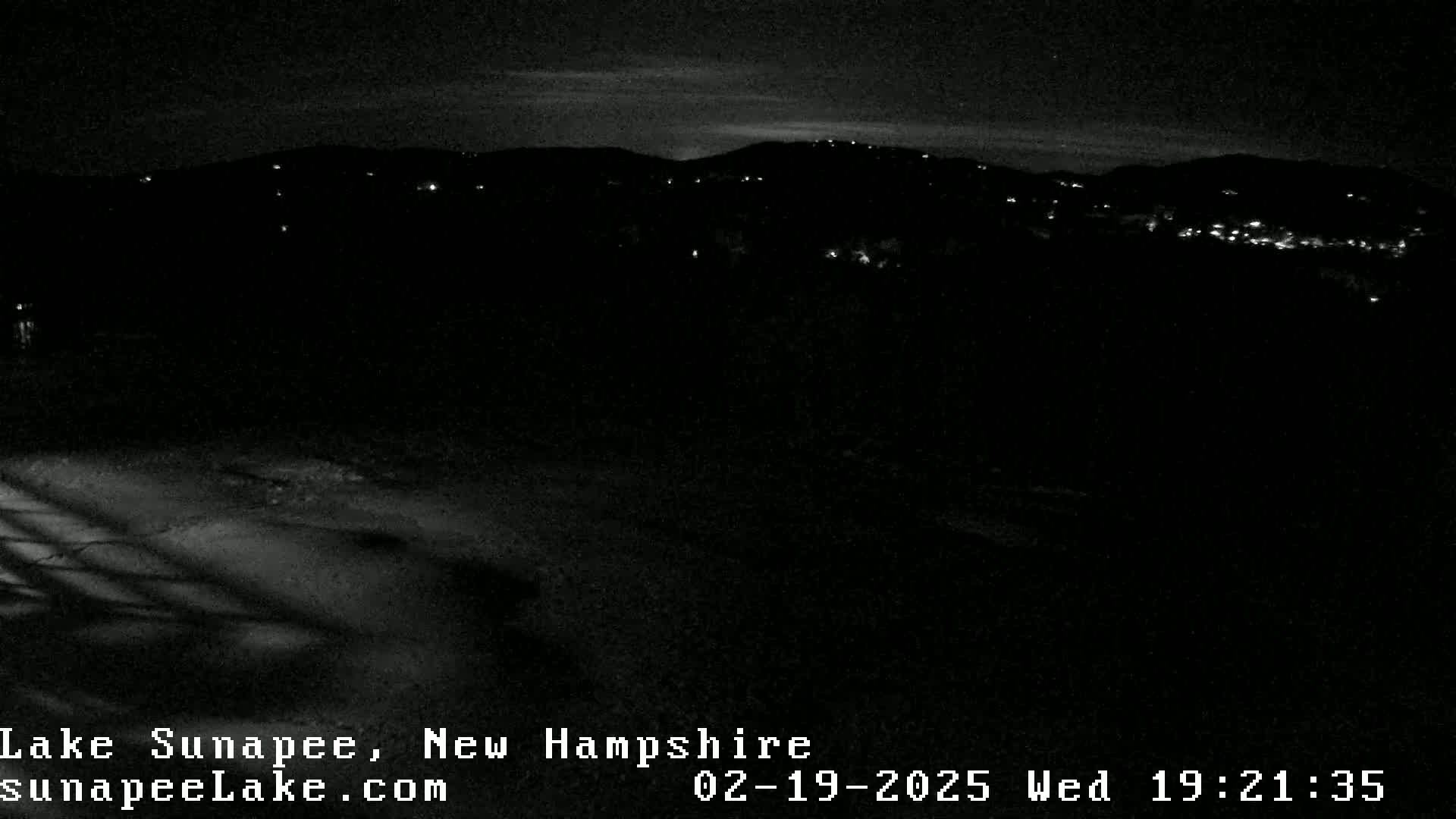A nighttime view shows a dark landscape with distant, sparsely lit hills under a mostly clear sky.
