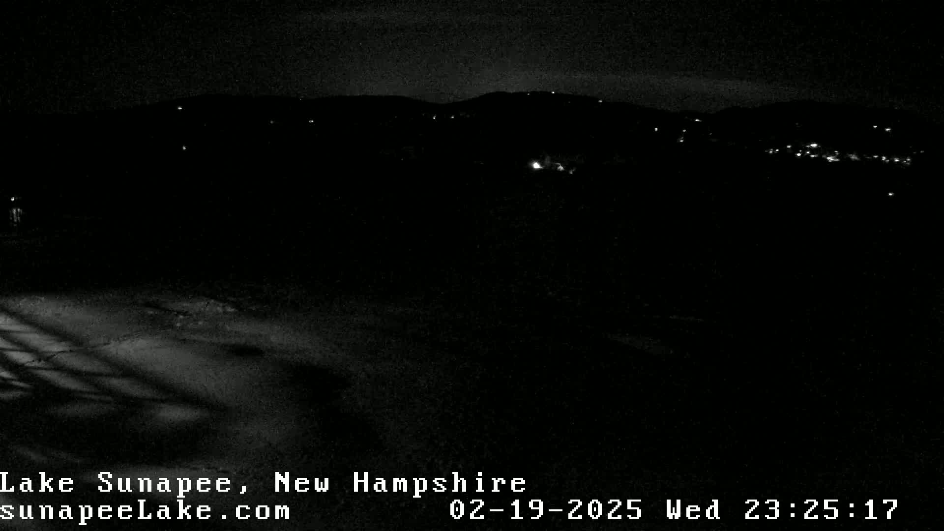 A nighttime, snow-covered landscape shows a dark hillside with faint lights in the distance.