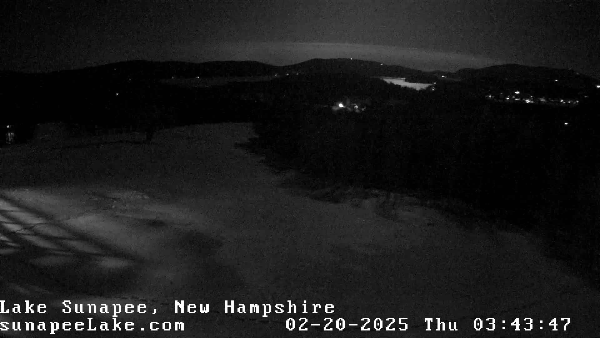 A nighttime view of a snow-covered landscape shows a dark, wooded area with a few distant lights under a clear, dark sky.