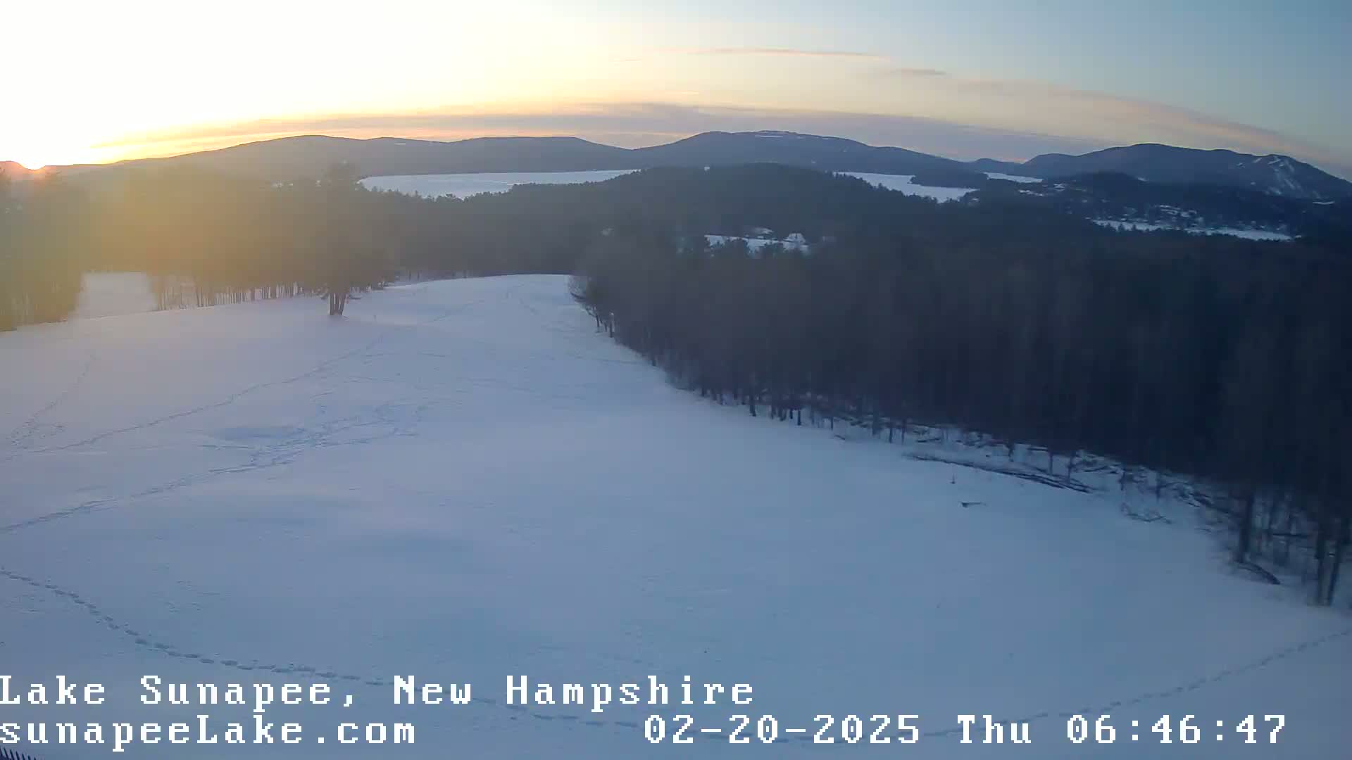 A snow-covered landscape at sunrise, with a forest and a lake visible in the distance.