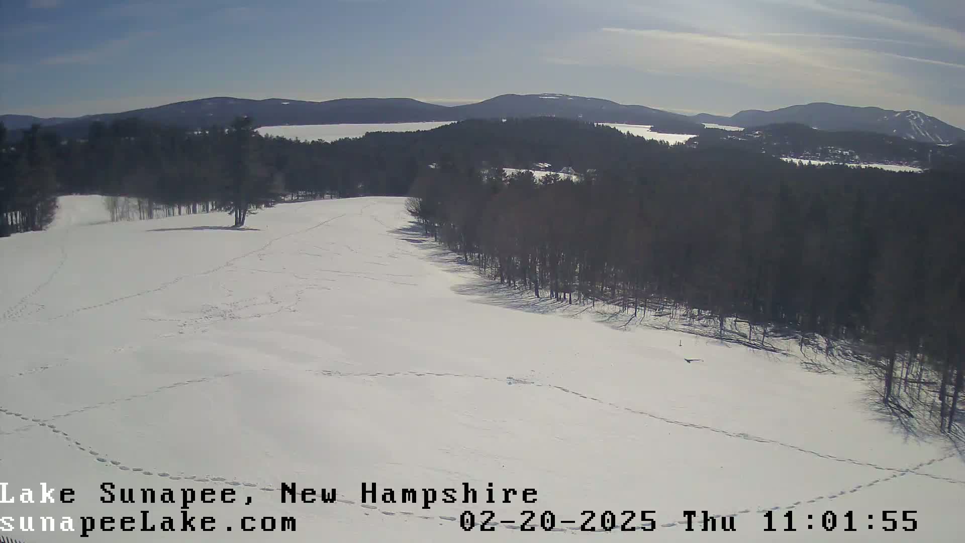 A snow-covered hill with footprints, overlooking a lake and distant mountains under a clear, sunny sky.