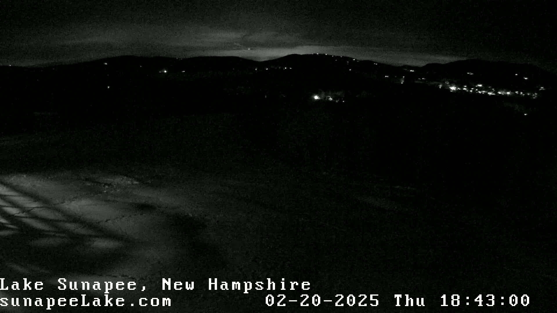 A nighttime view of dimly lit distant hills and a dark foreground under a mostly clear sky.