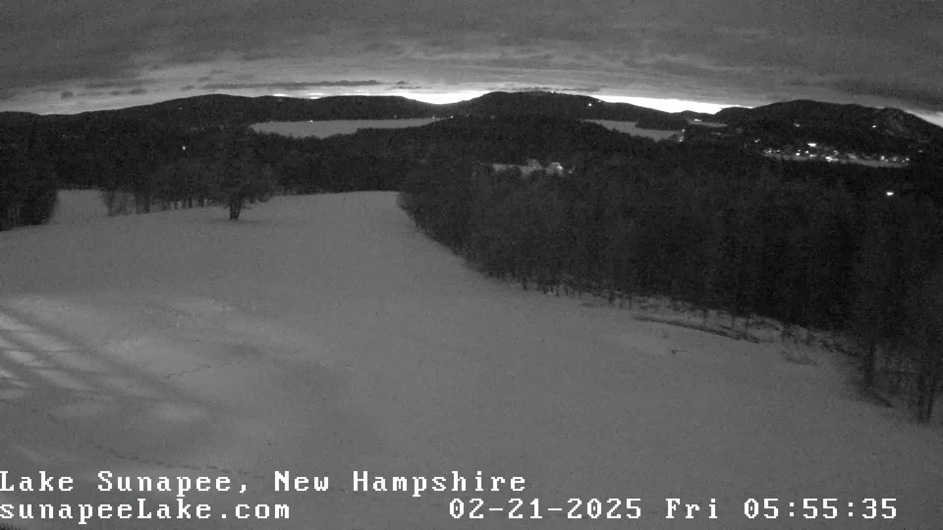 A snow-covered landscape at twilight shows a hilltop with a dark forest in the background under a cloudy sky.