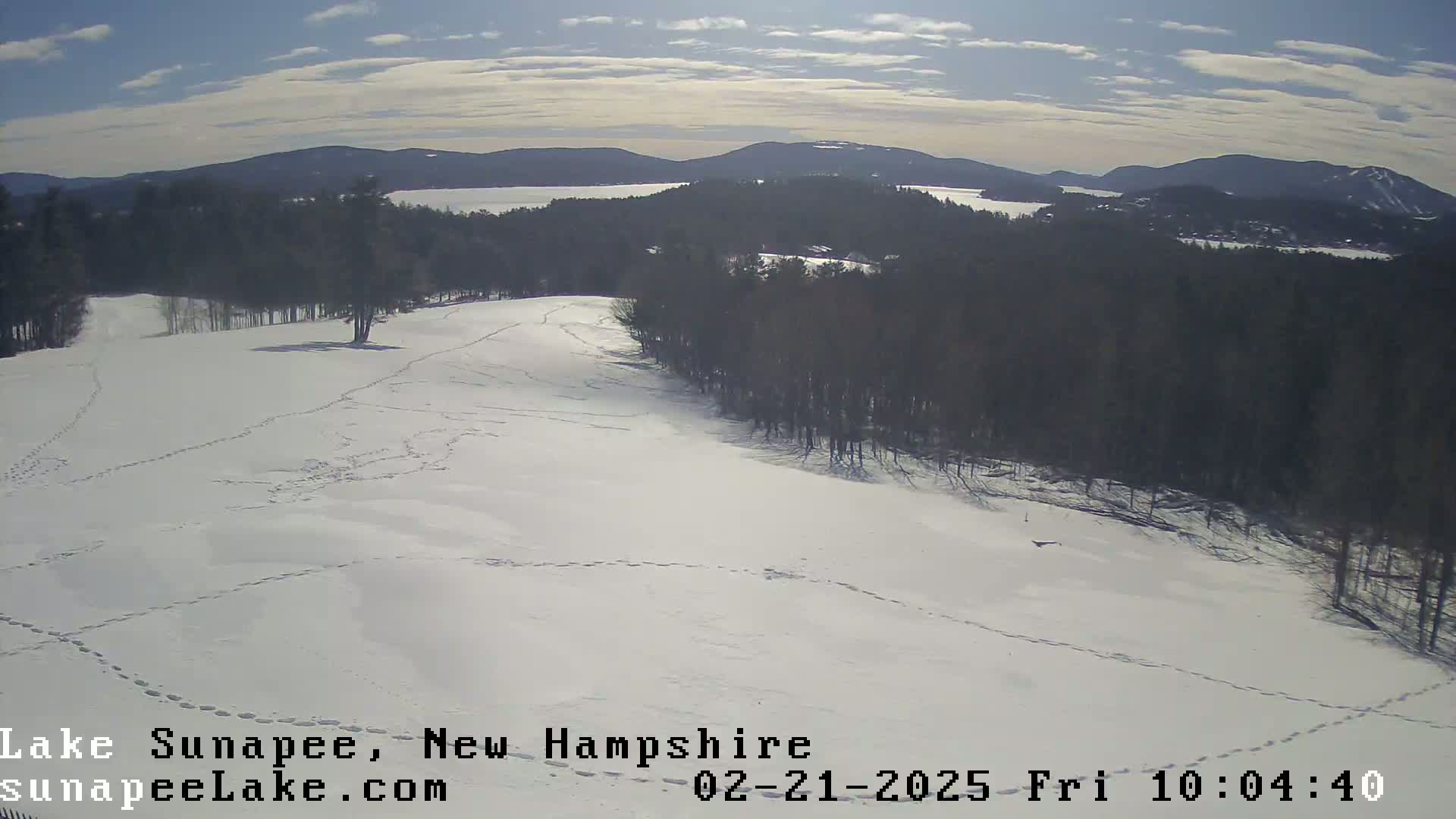 A snow-covered field with footprints, overlooking a lake and distant mountains under a partly cloudy sky.