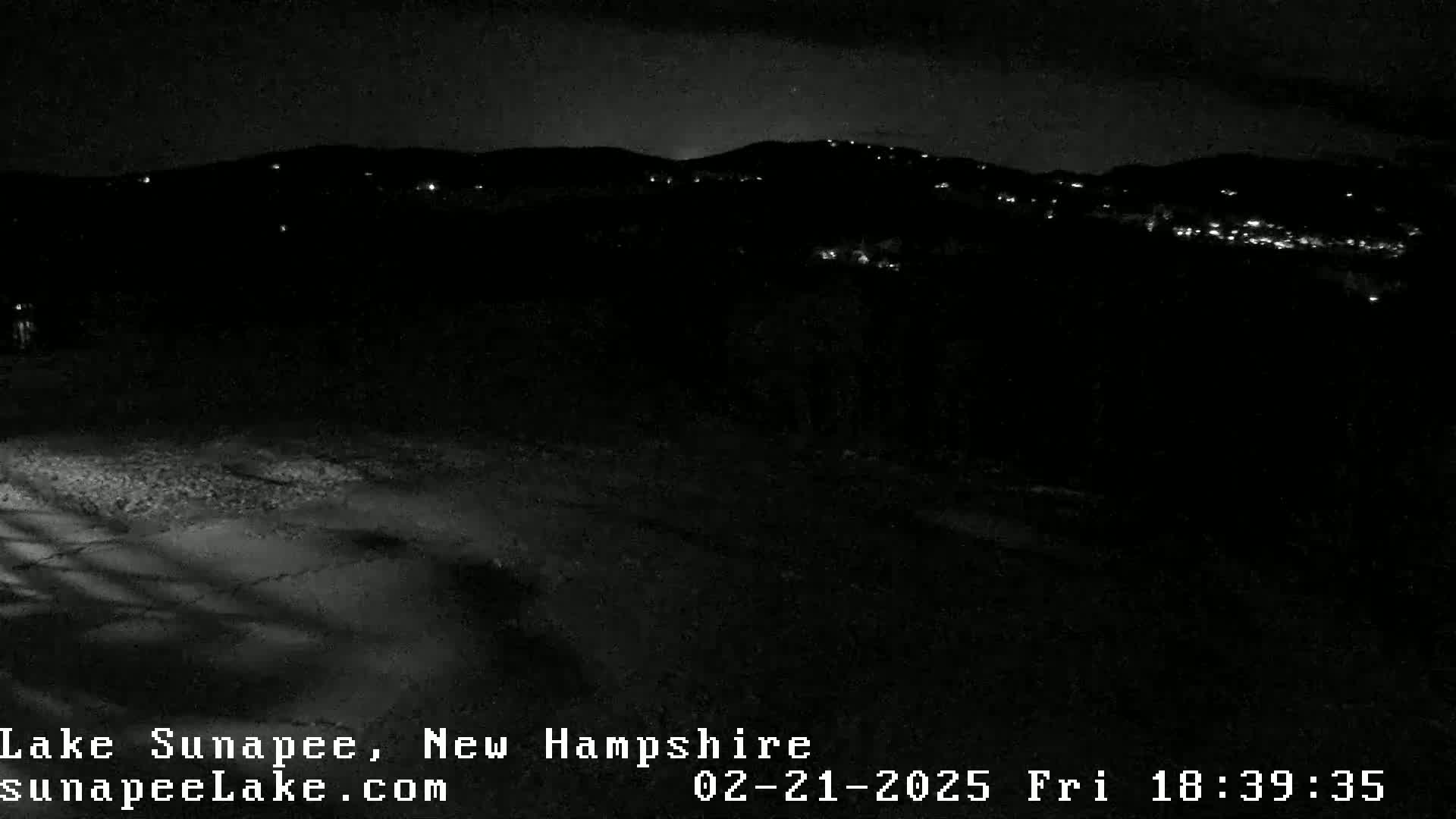 A nighttime view of a snow-covered area in front of a dark, hilly landscape with scattered lights.