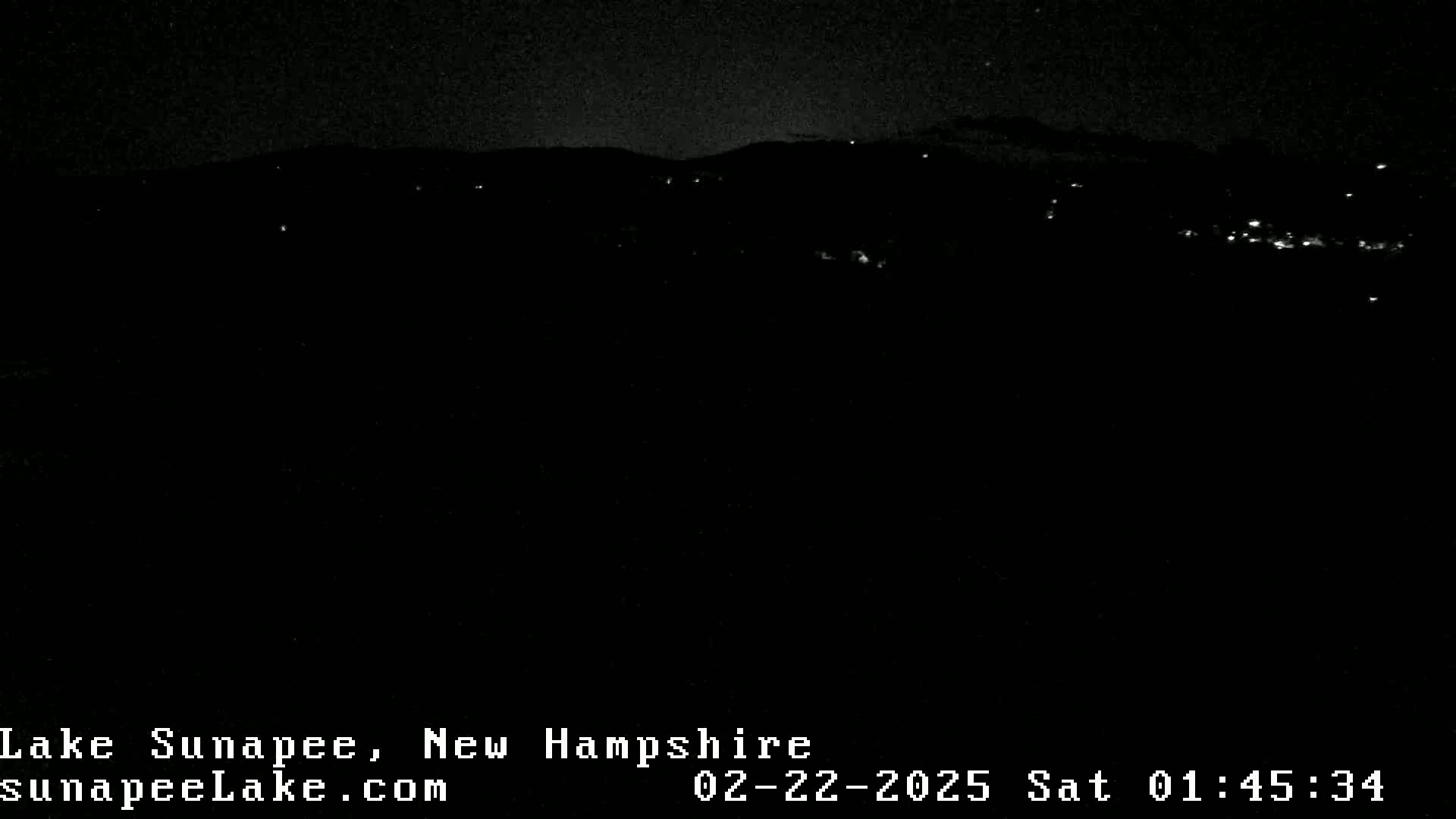 A dark nighttime landscape shows a distant line of faint lights across a hilly horizon under a clear sky.