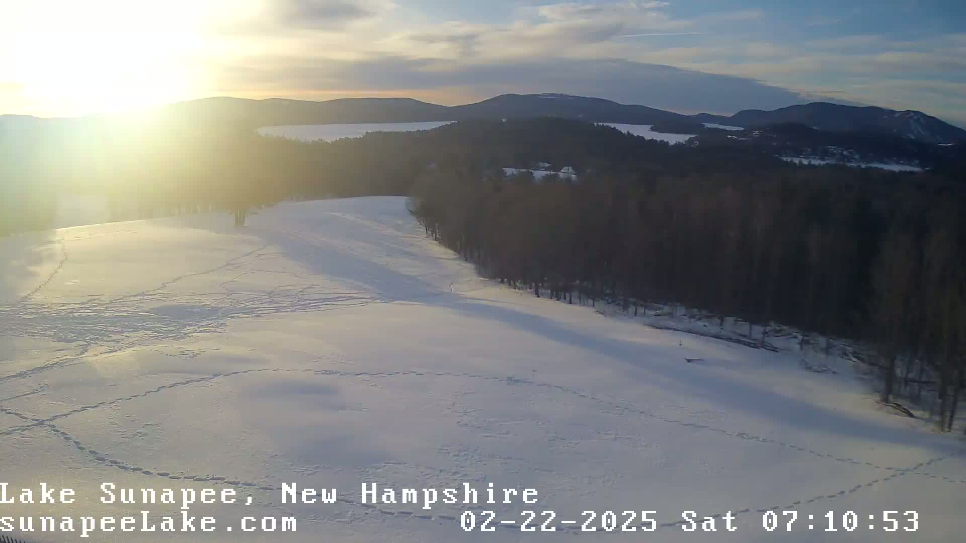 A snow-covered landscape with footprints and a distant lake is seen at sunrise.