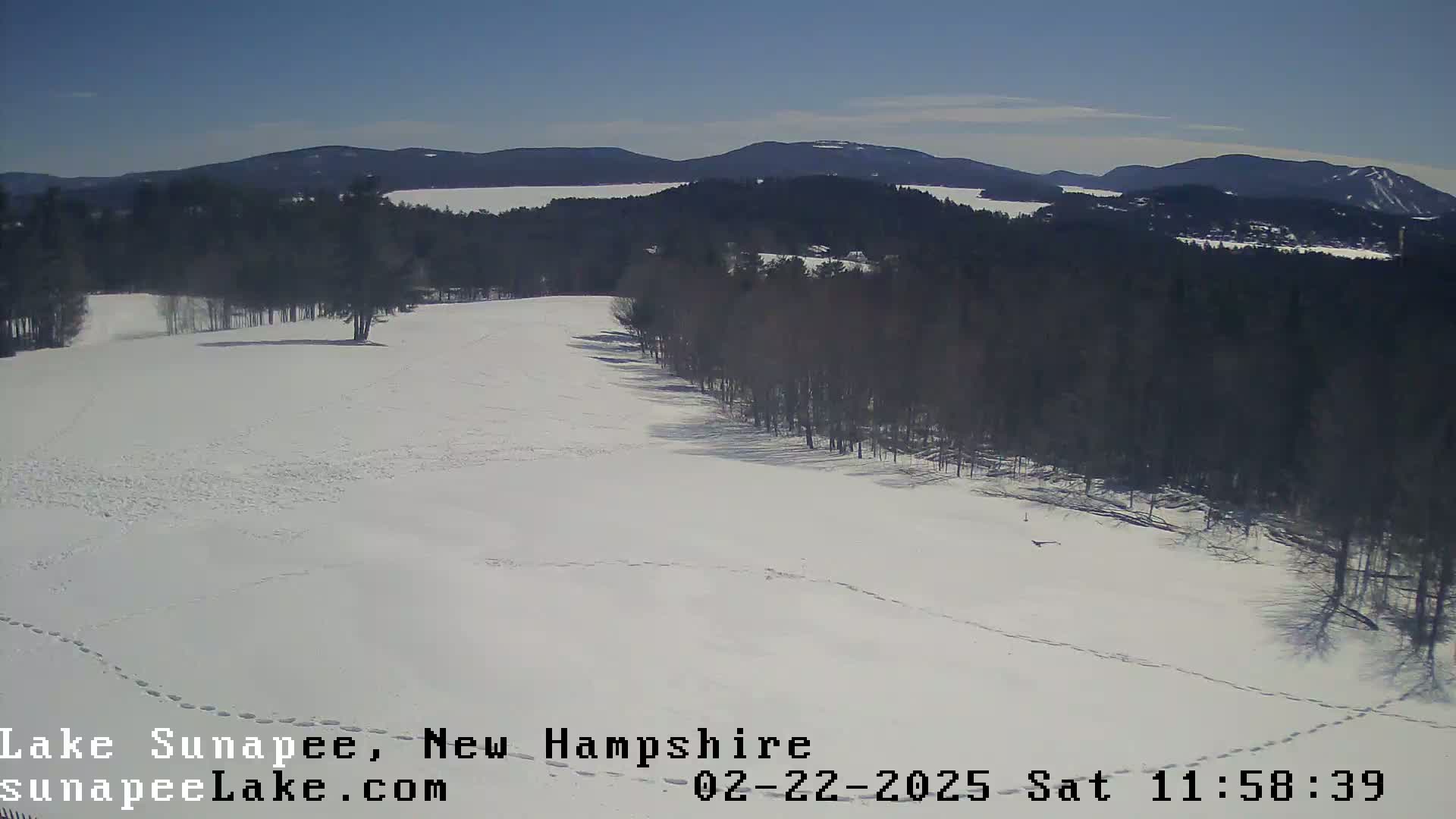 A snow-covered field with footprints, bordered by a line of leafless trees, overlooks a lake and distant mountains under a clear, sunny sky.