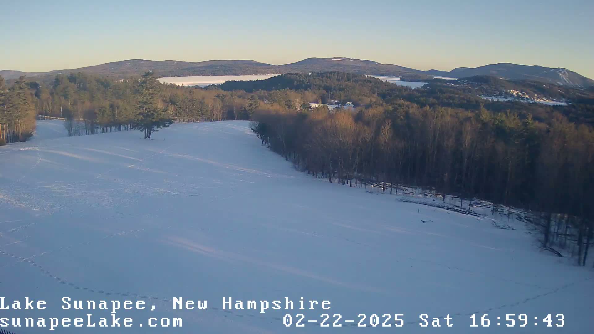 A snow-covered hillside slopes down towards a lake and distant mountains under a clear, light-blue sky.