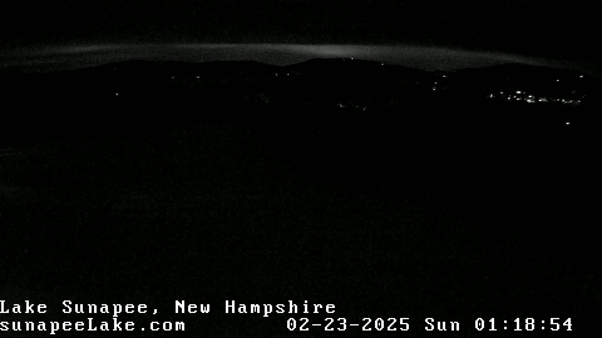 A nighttime long shot shows a dark landscape with faintly visible lights from a distant town, under a mostly clear sky.
