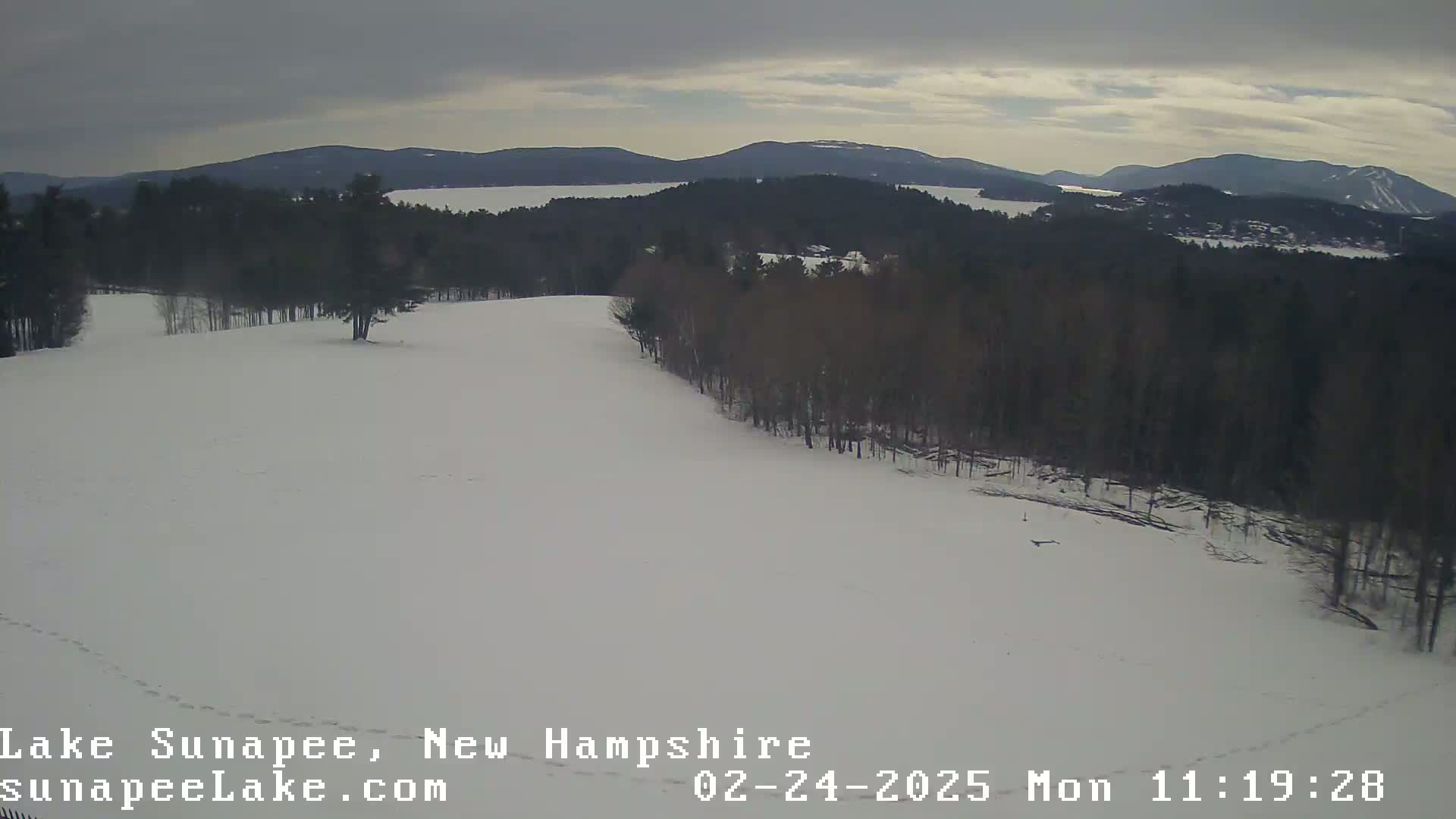A snow-covered landscape shows a distant lake and mountains under an overcast sky.