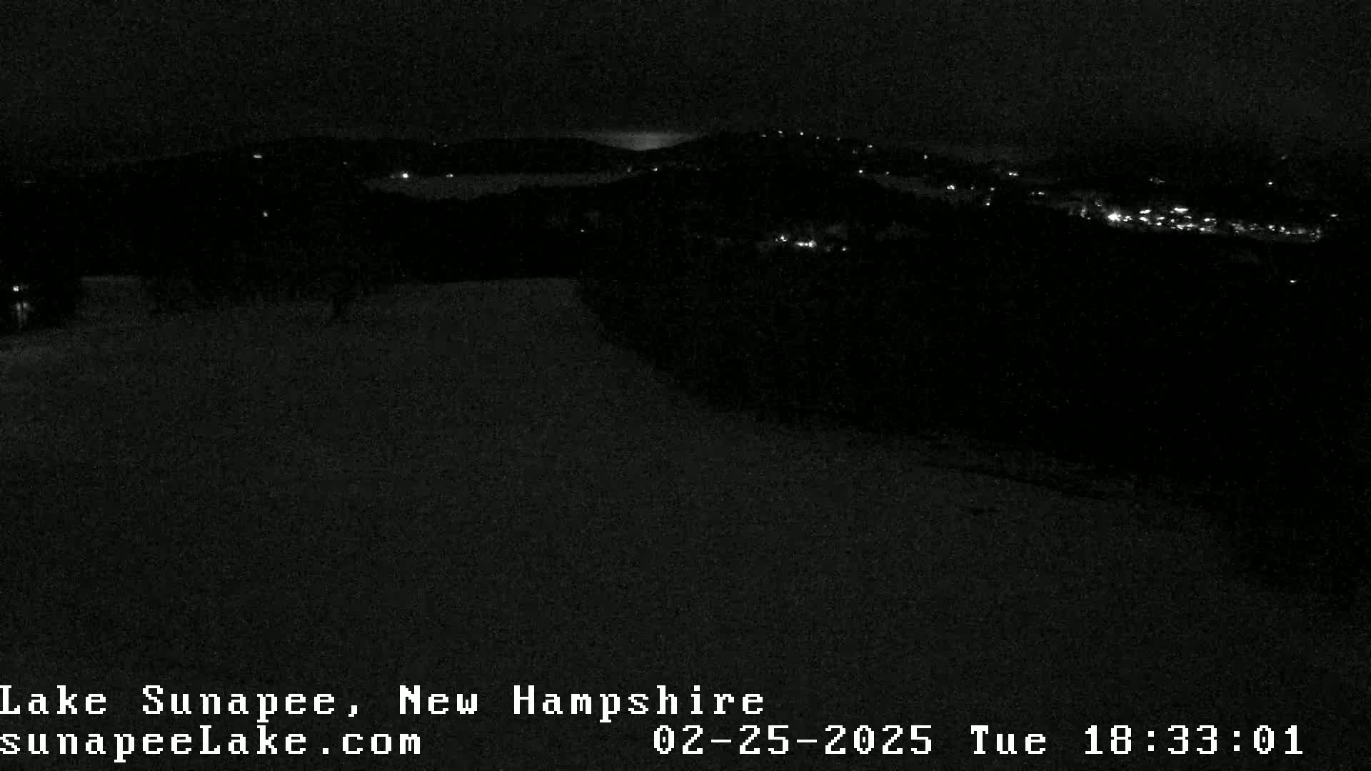 A nighttime, snow-covered landscape is dimly lit by scattered lights from a distant town under a dark sky.