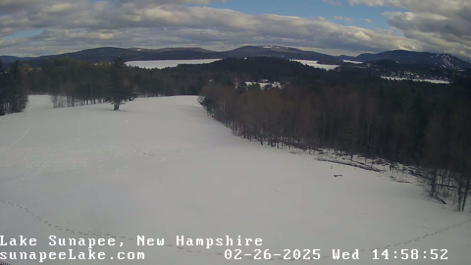 A snow-covered field with animal tracks, bordered by a dark forest, overlooks a lake and distant mountains under a partly cloudy sky.