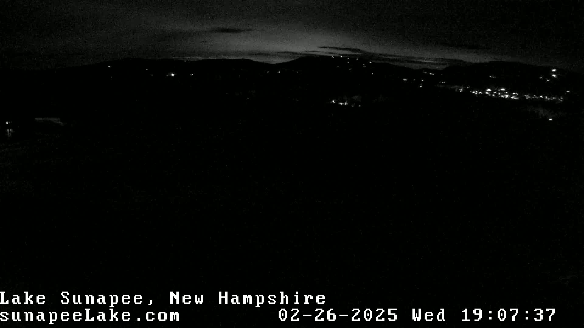 A nighttime, long shot of a dark landscape with a few distant lights visible on a hillside under a mostly clear sky.