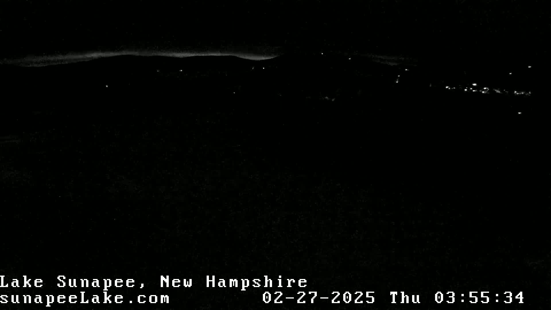A nighttime view of a dark landscape with a few distant lights and a faint, light horizon.