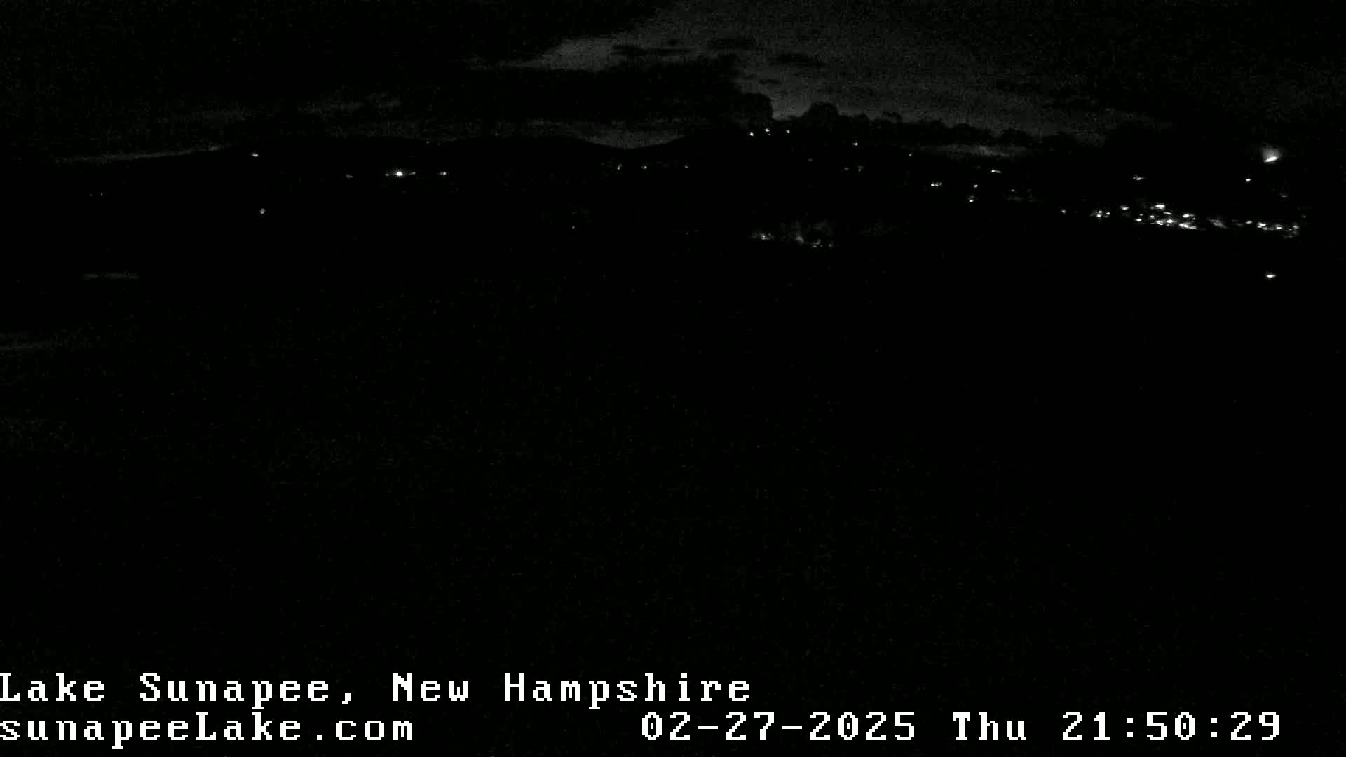 A nighttime, long-range view of a dimly lit town nestled among dark hills under a cloudy sky.