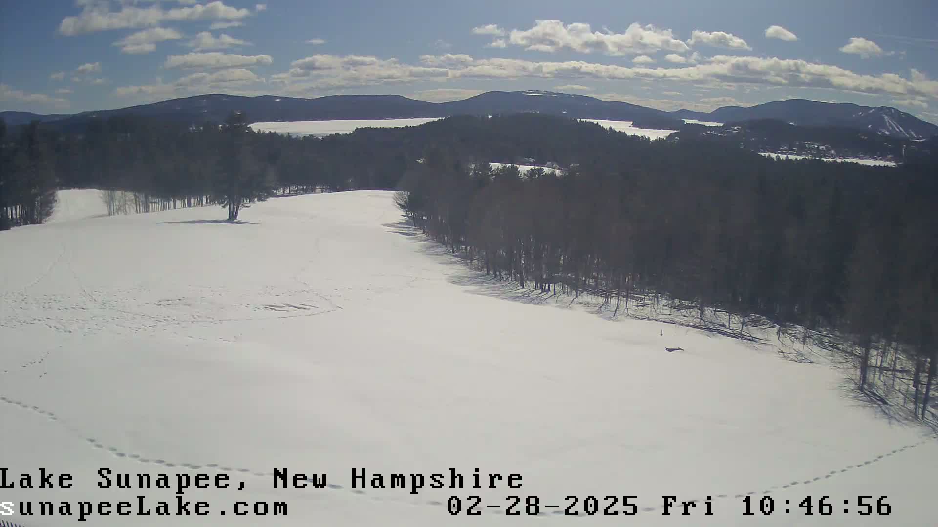 A snow-covered landscape with a line of trees bordering a partially visible lake under a partly sunny sky.