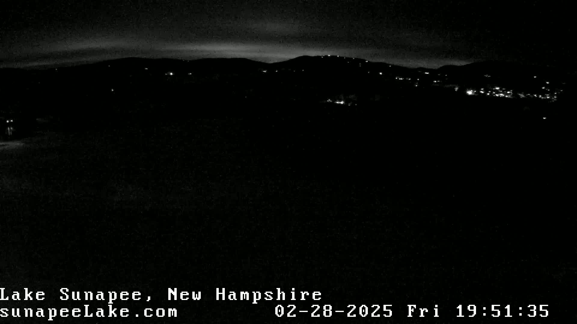 A nighttime, dark landscape shows a distant line of lights nestled in the hills under a mostly clear sky.
