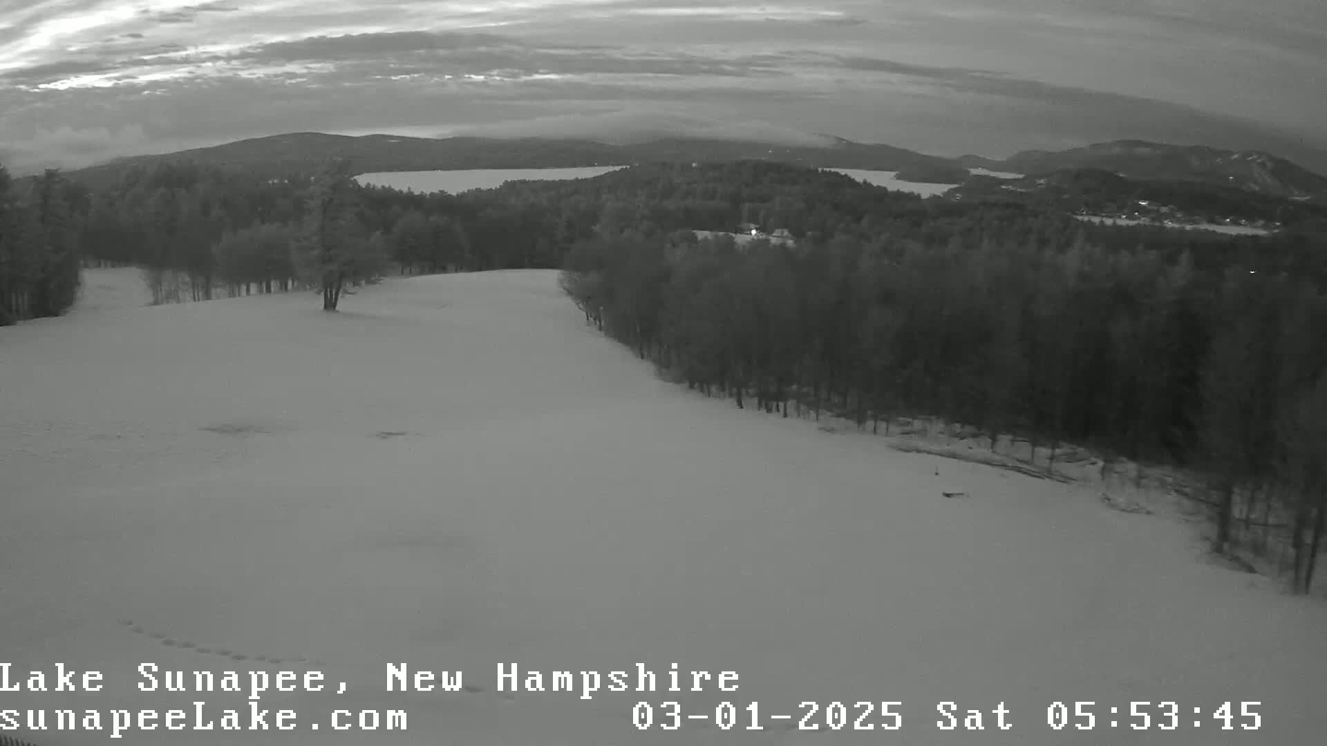 A snowy, lightly wooded hill overlooks a calm lake under a cloudy, twilight sky.