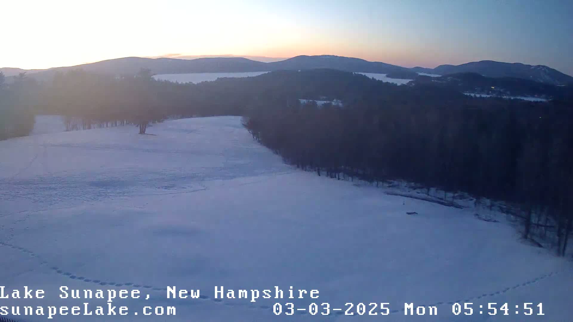 A snow-covered landscape at sunrise shows a  wooded area overlooking a lake and distant mountains.
