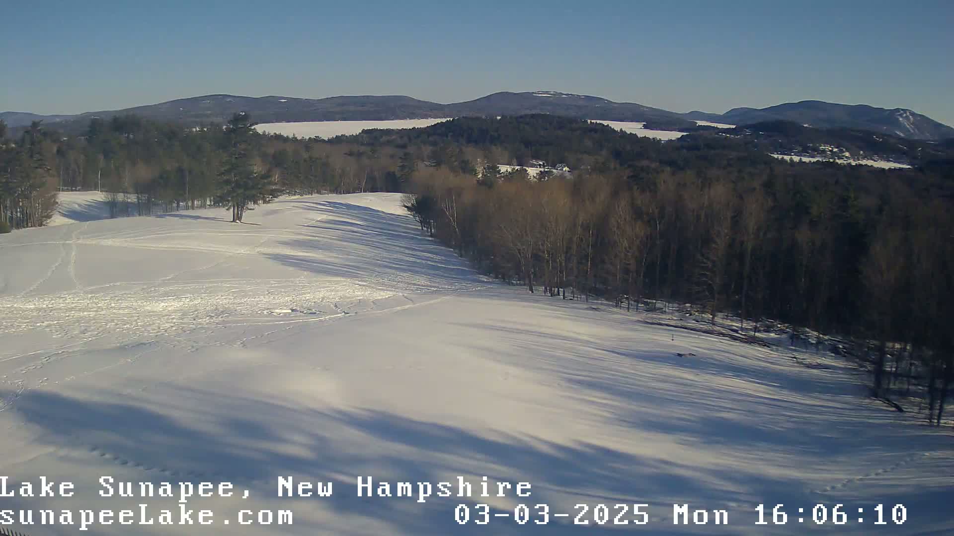 A snow-covered landscape with a line of trees and distant mountains under a clear, sunny sky.