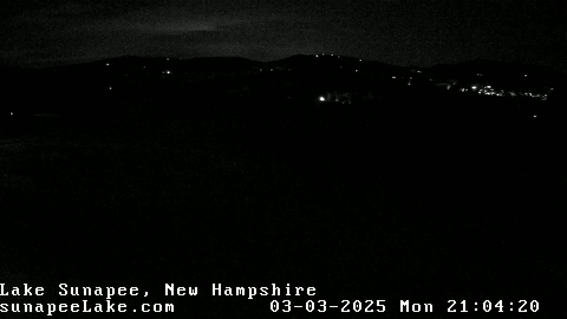 A nighttime, dark view of a distant hilltop with a few scattered lights visible under a mostly clear sky.