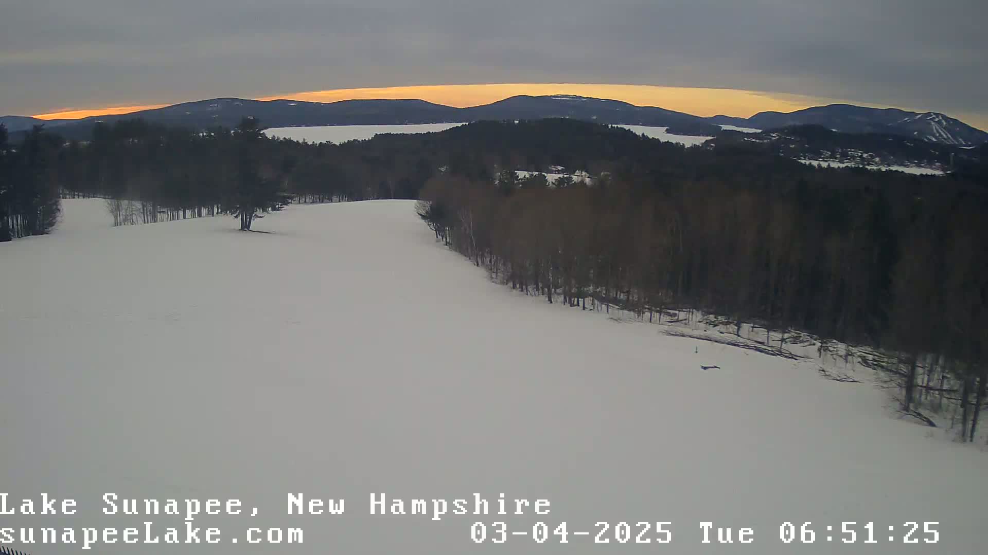 A snow-covered landscape with a line of trees bordering a wide, open snowfield, overlooking a lake and distant mountains under a dusky sky.