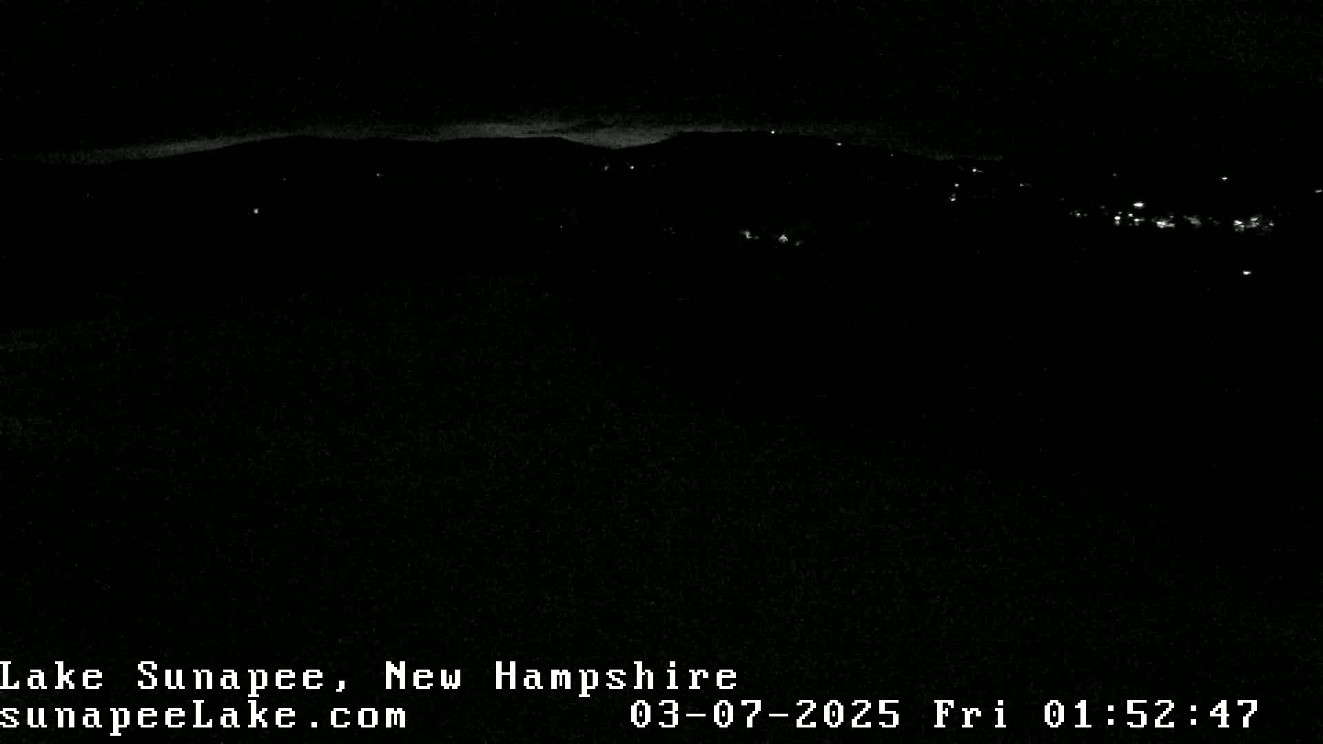 A nighttime long shot of a dark, hilly landscape with a few distant lights visible under a clear sky.