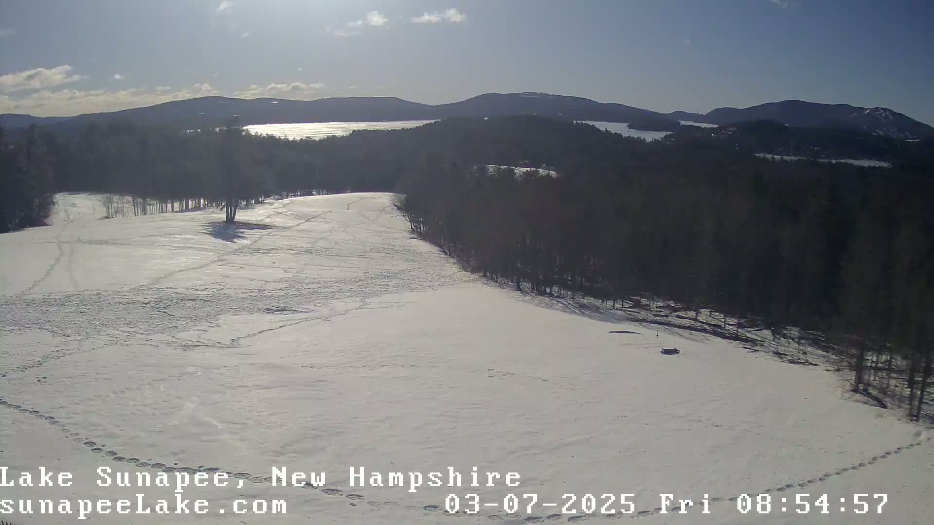A snow-covered field with footprints and a distant view of a lake and mountains under a mostly sunny sky.
