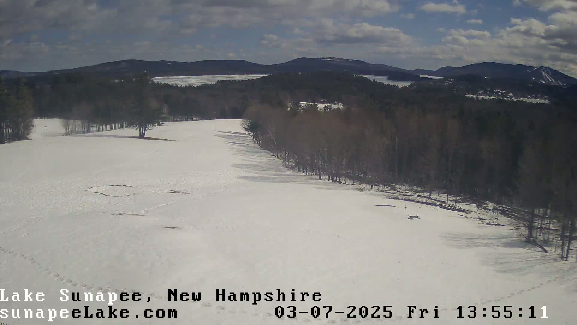 A snow-covered hillside slopes down toward a wooded area and a distant lake under a partly cloudy sky.