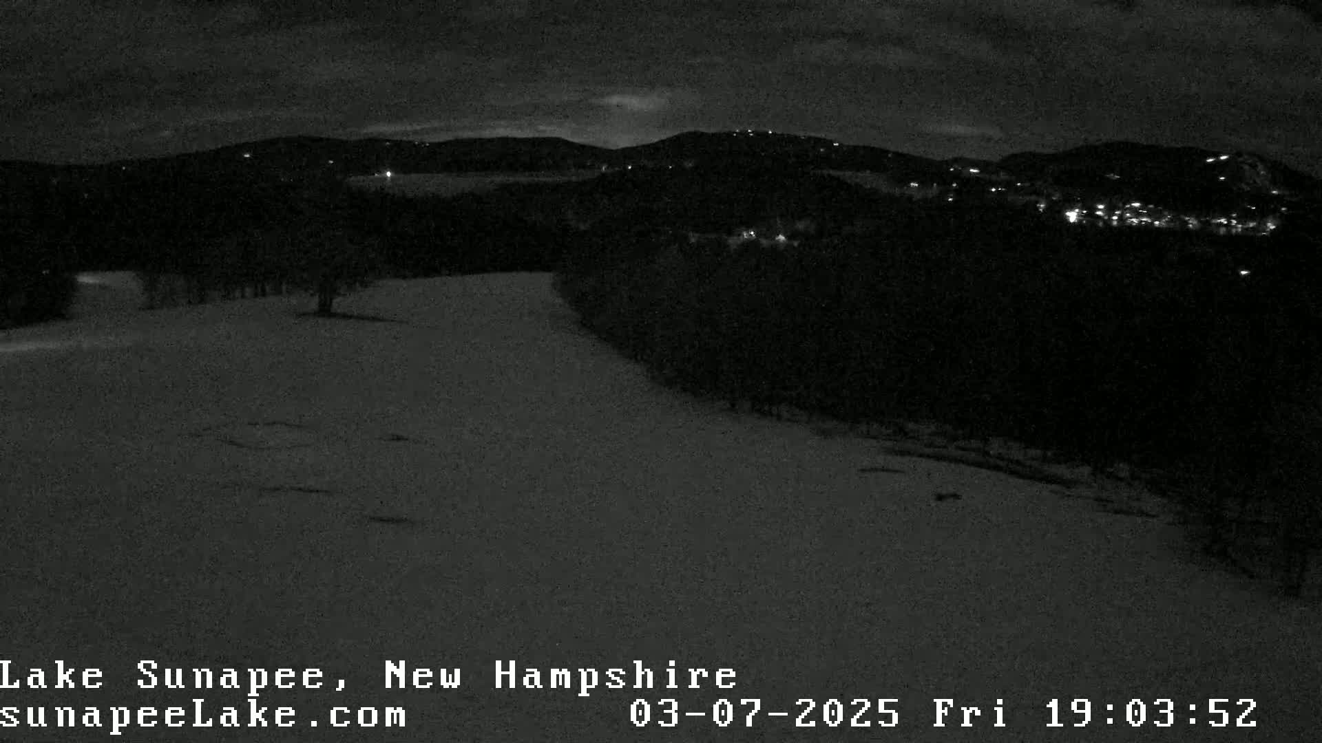 A snow-covered landscape at night is illuminated by distant lights from a town nestled in the hills.