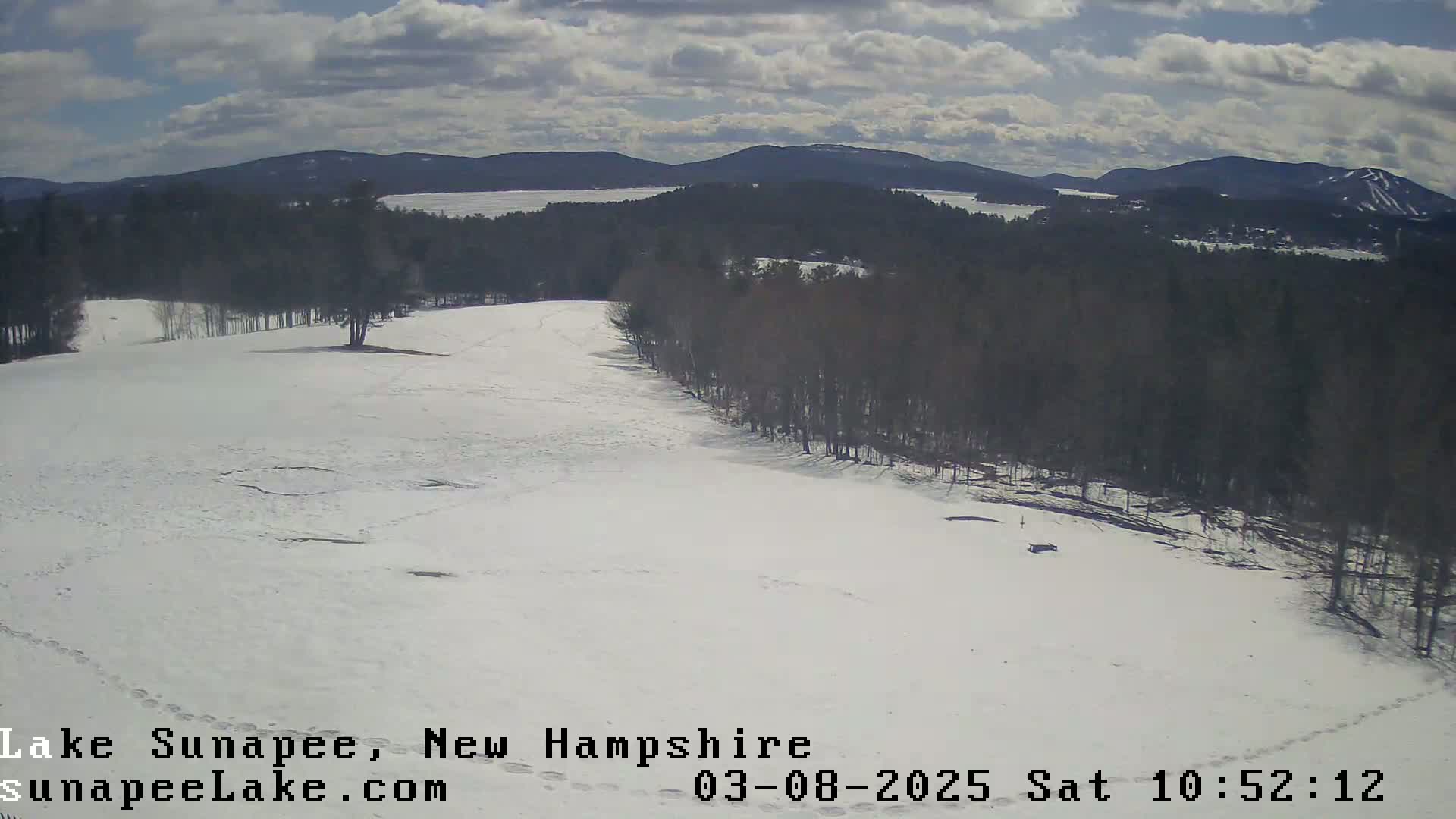 A snow-covered landscape with a line of trees to the right and mountains in the distance under a partly cloudy sky.
