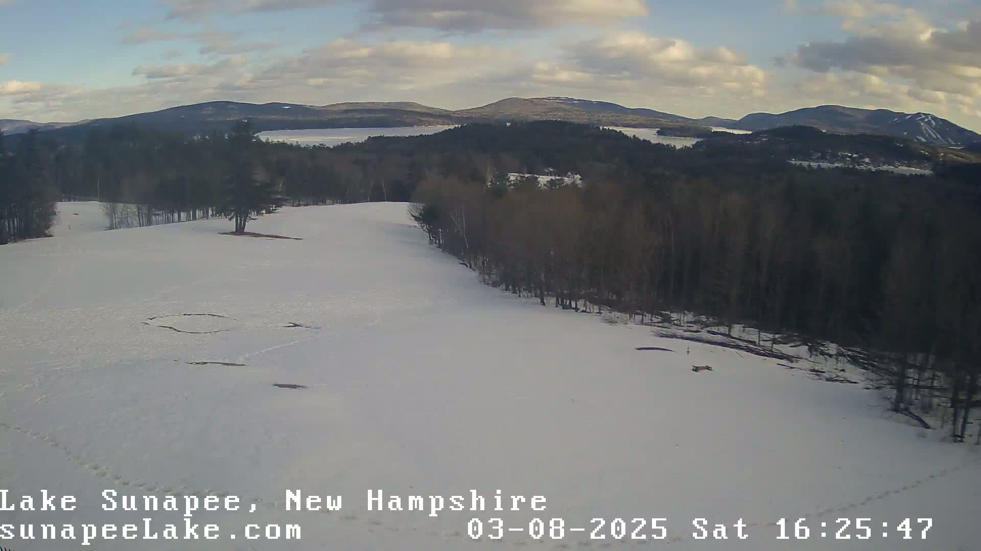 A snow-covered landscape with a distant view of a lake and mountains under a partly cloudy sky.
