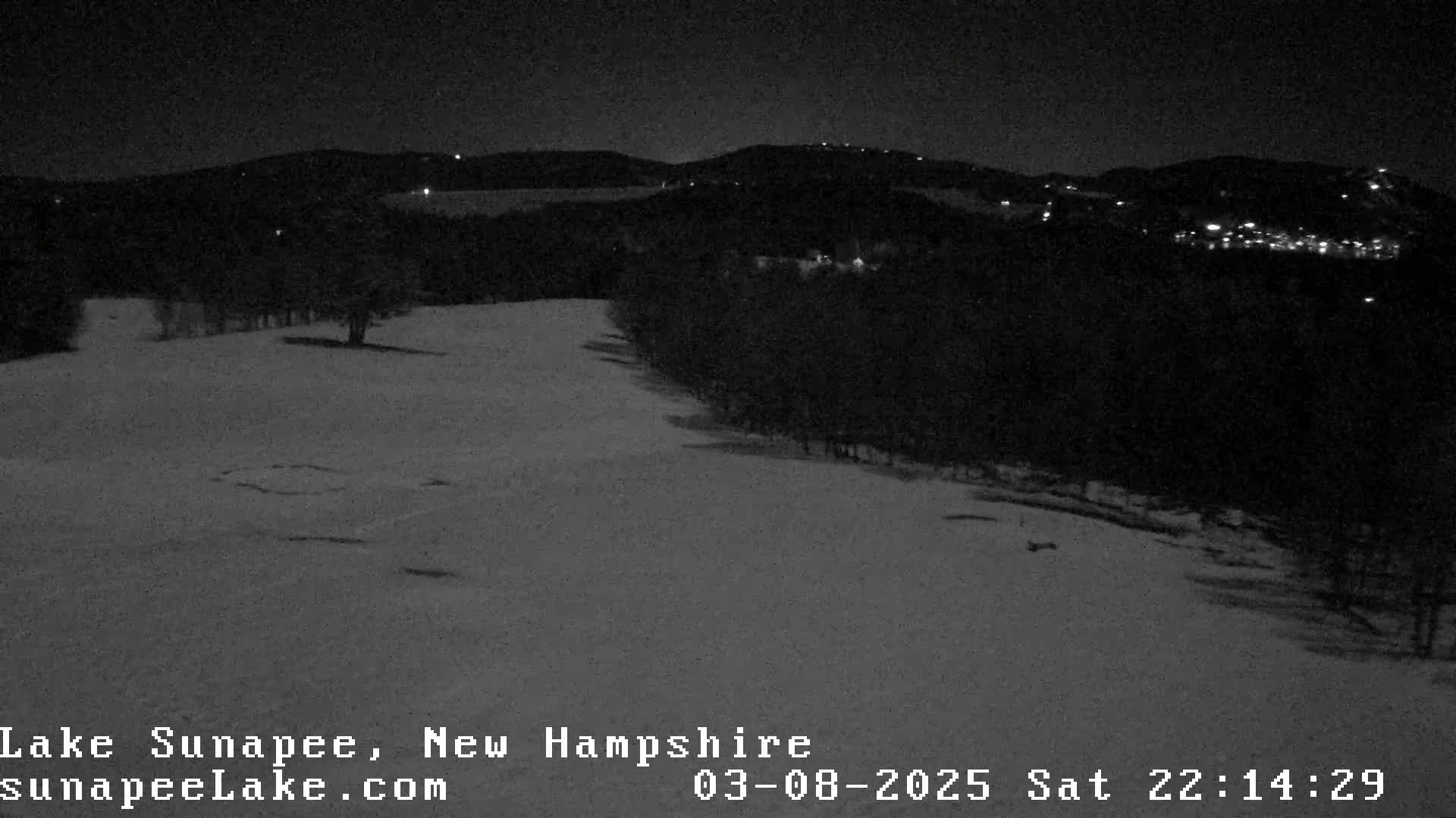 A snow-covered landscape at night, with distant lights illuminating a tree line and hills.