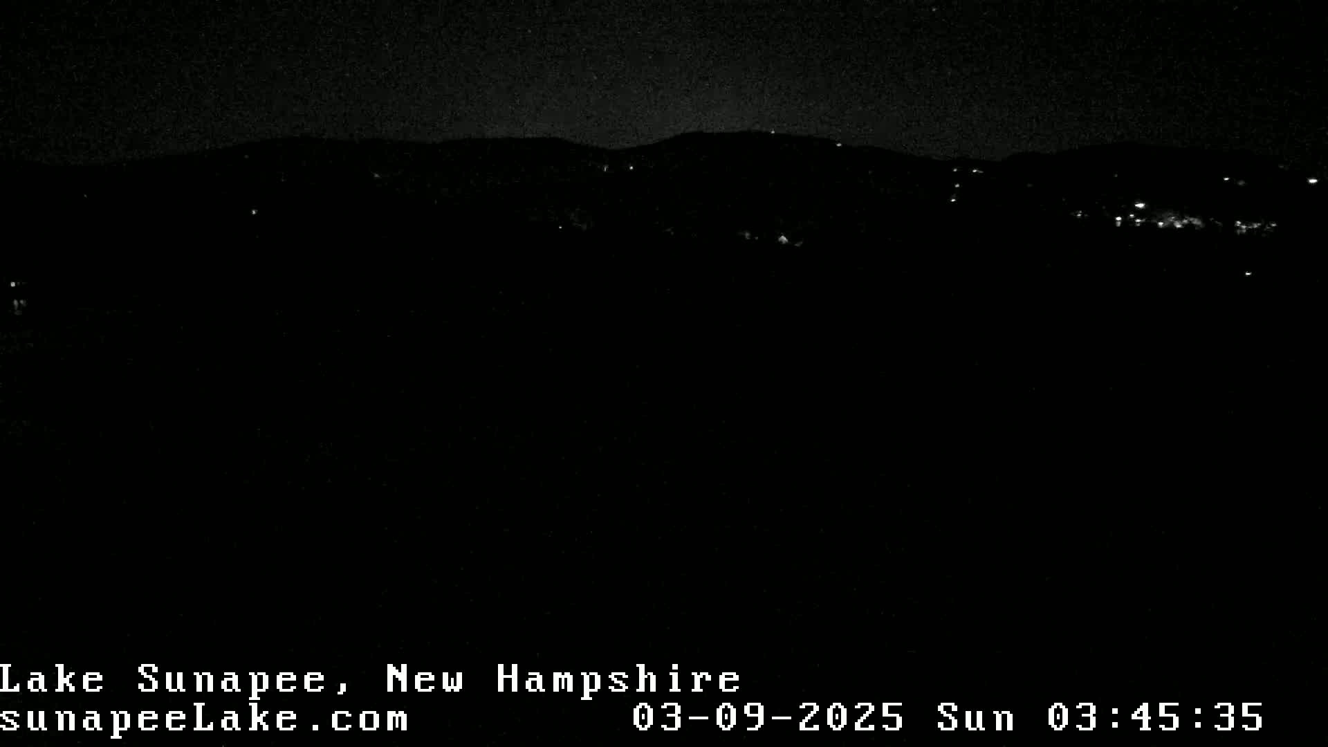 A nighttime, dark landscape shows a distant line of lights and hills under a clear, starry sky.
