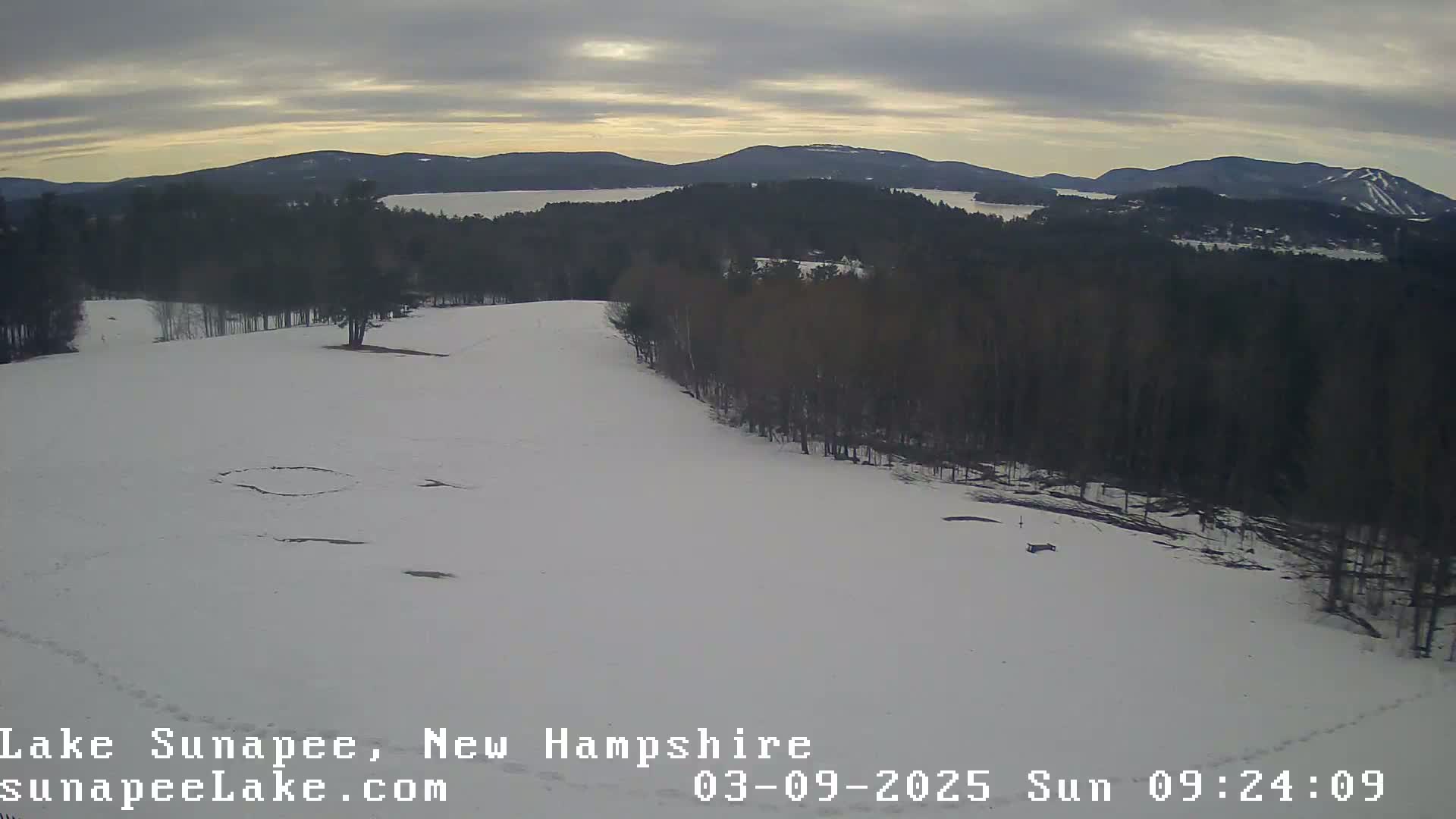 A snow-covered landscape with a view of a lake and distant mountains under an overcast sky.