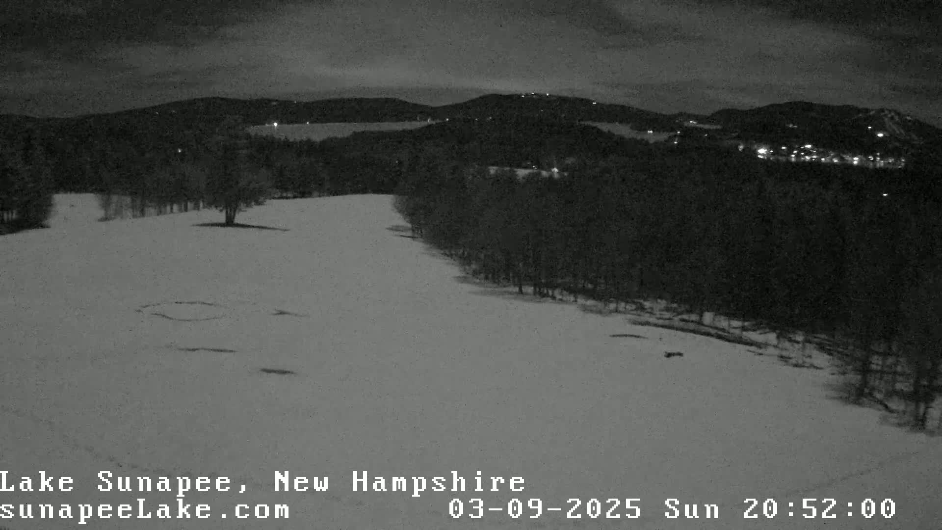 A snow-covered landscape at night, showing a hilly area with dark trees and distant lights under a dark sky.