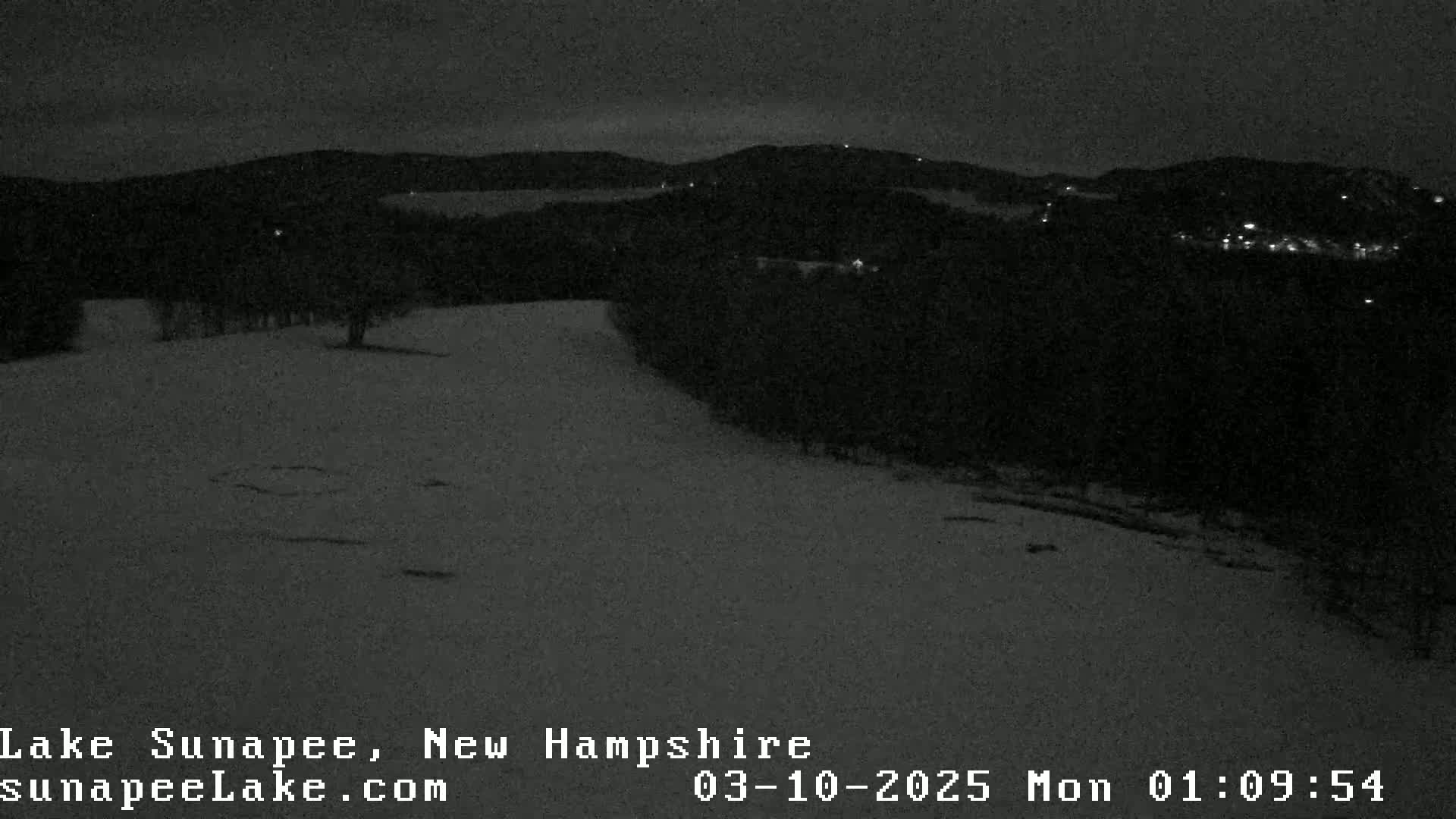 A nighttime, snow-covered landscape shows dark hills in the distance, illuminated by a few faint lights.