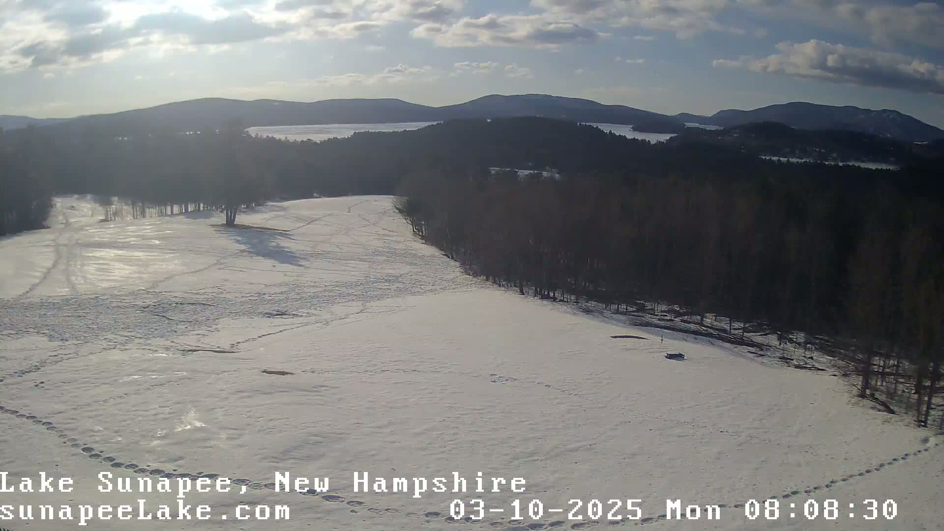 A snow-covered field with footprints and a line of trees in the background, under a partly sunny sky.