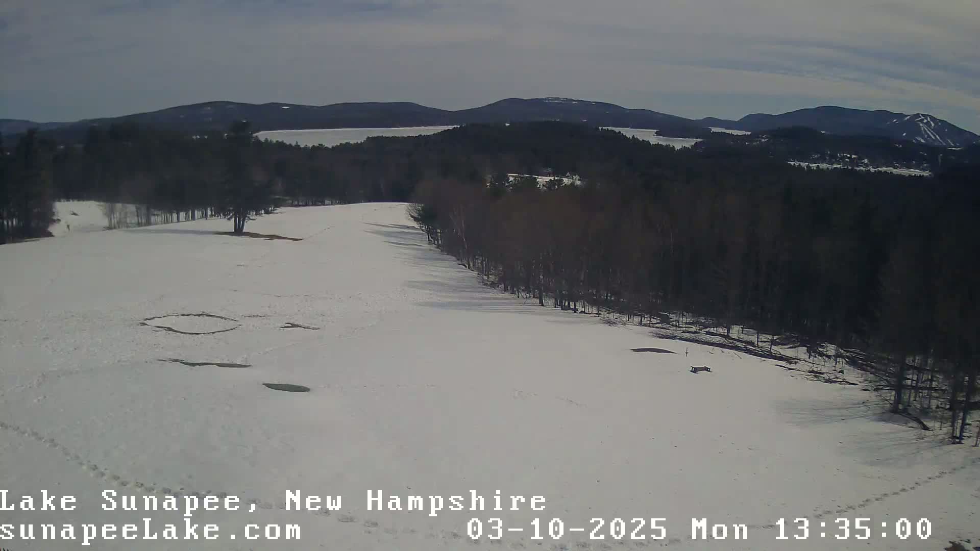 A snow-covered landscape with a line of bare trees separating a snow-covered field from a lake and distant mountains under a partly cloudy sky.