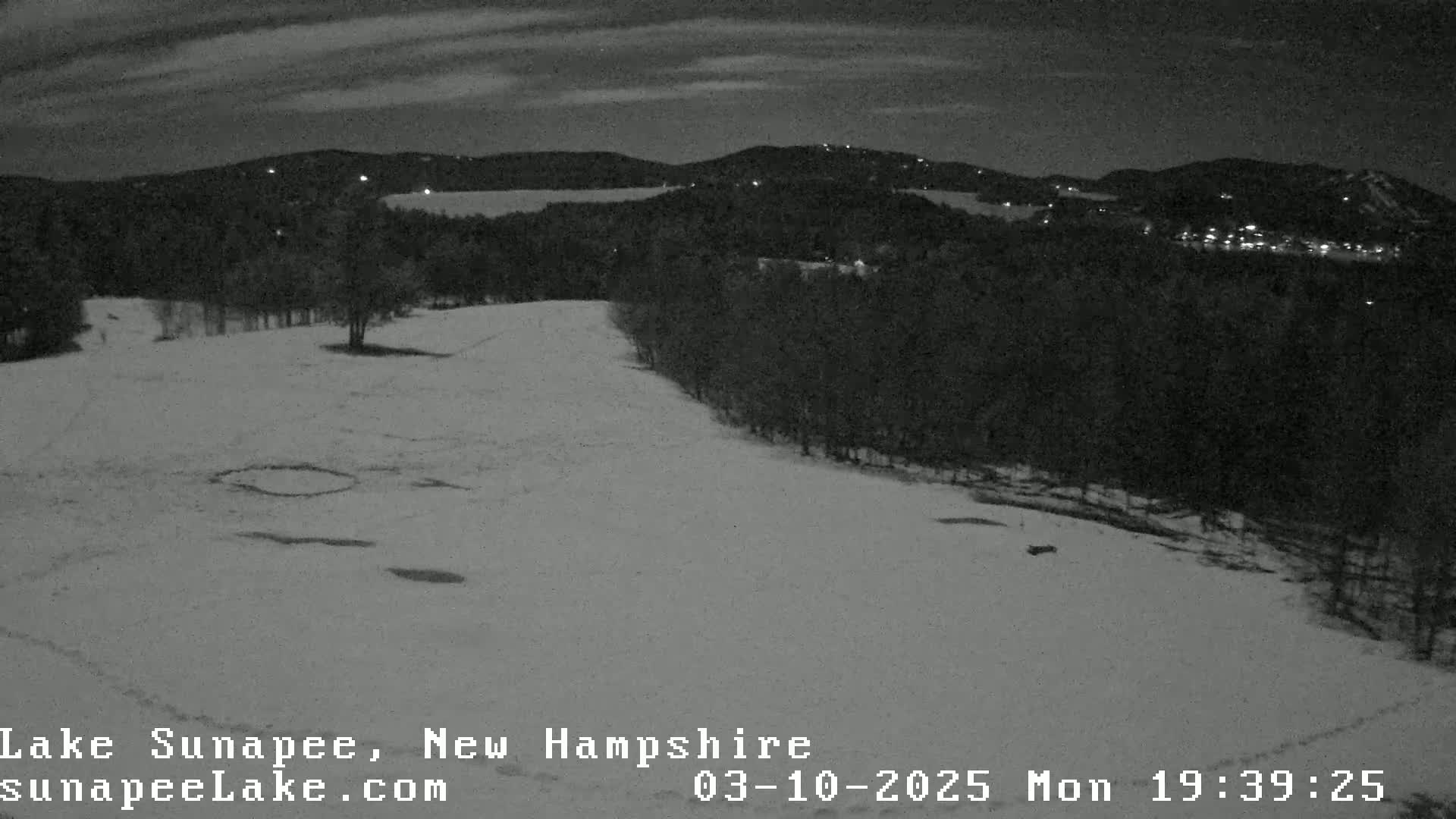 A snow-covered hill at night, overlooking a dark, wooded area with distant lights.